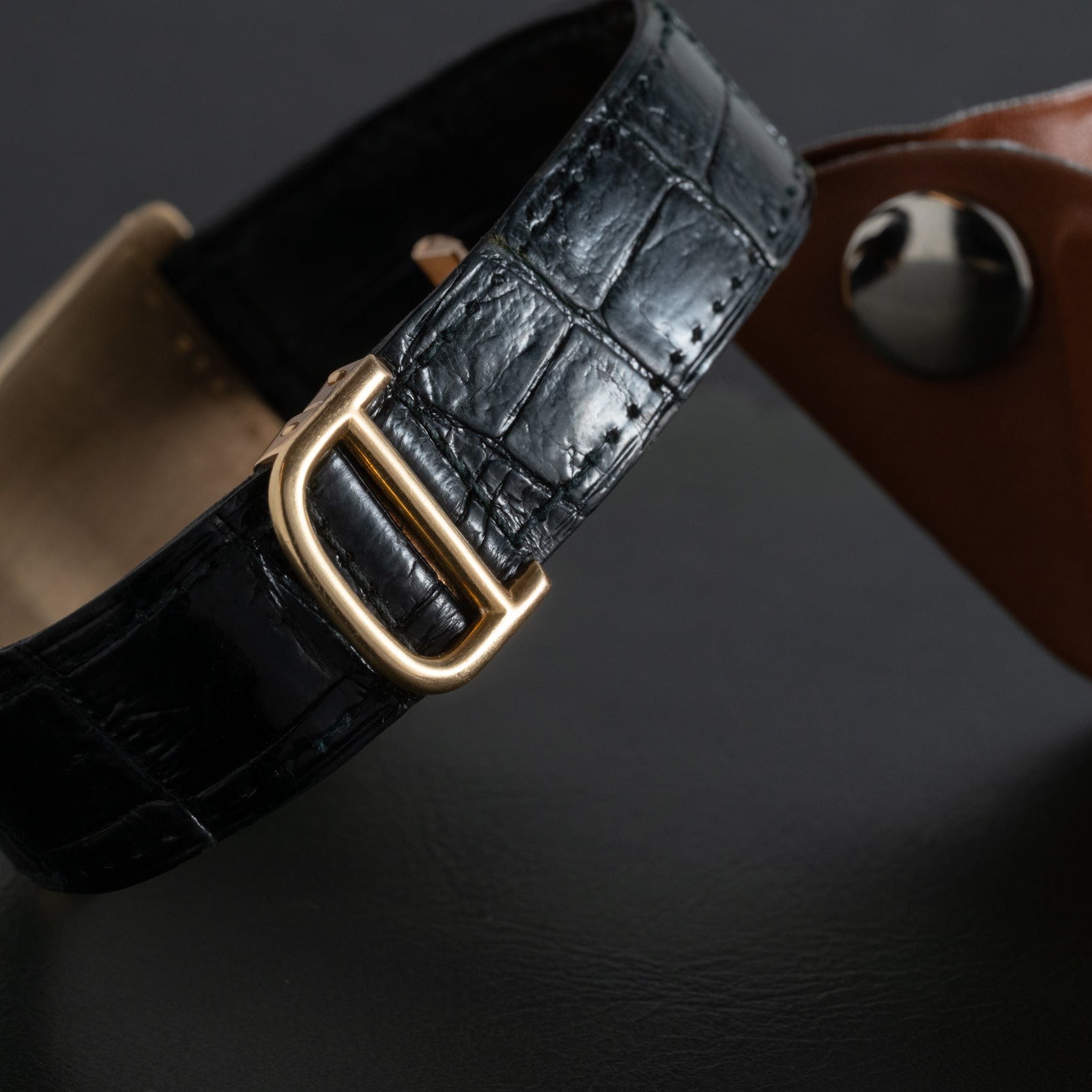 Close-up of the Cartier Tank Cintrée Jumbo black leather watch strap with a gold buckle, placed beside a brown leather item on a dark surface.