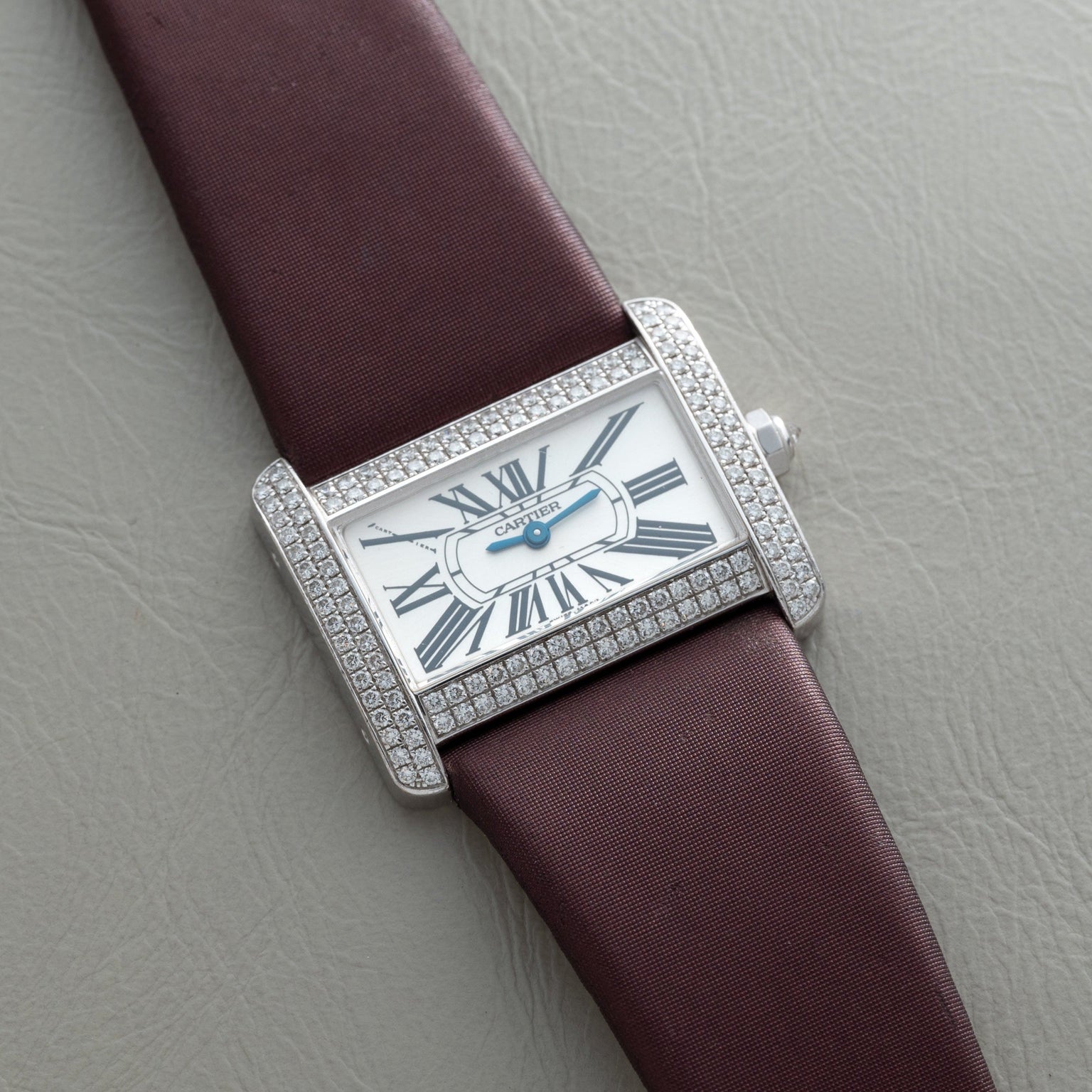 A Cartier Tank Divan watch with a diamond bezel, Roman numerals, and a brown strap is displayed on a gray surface.