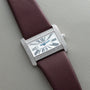 A Cartier Tank Divan watch with a diamond bezel, Roman numerals, and a brown strap is displayed on a gray surface.