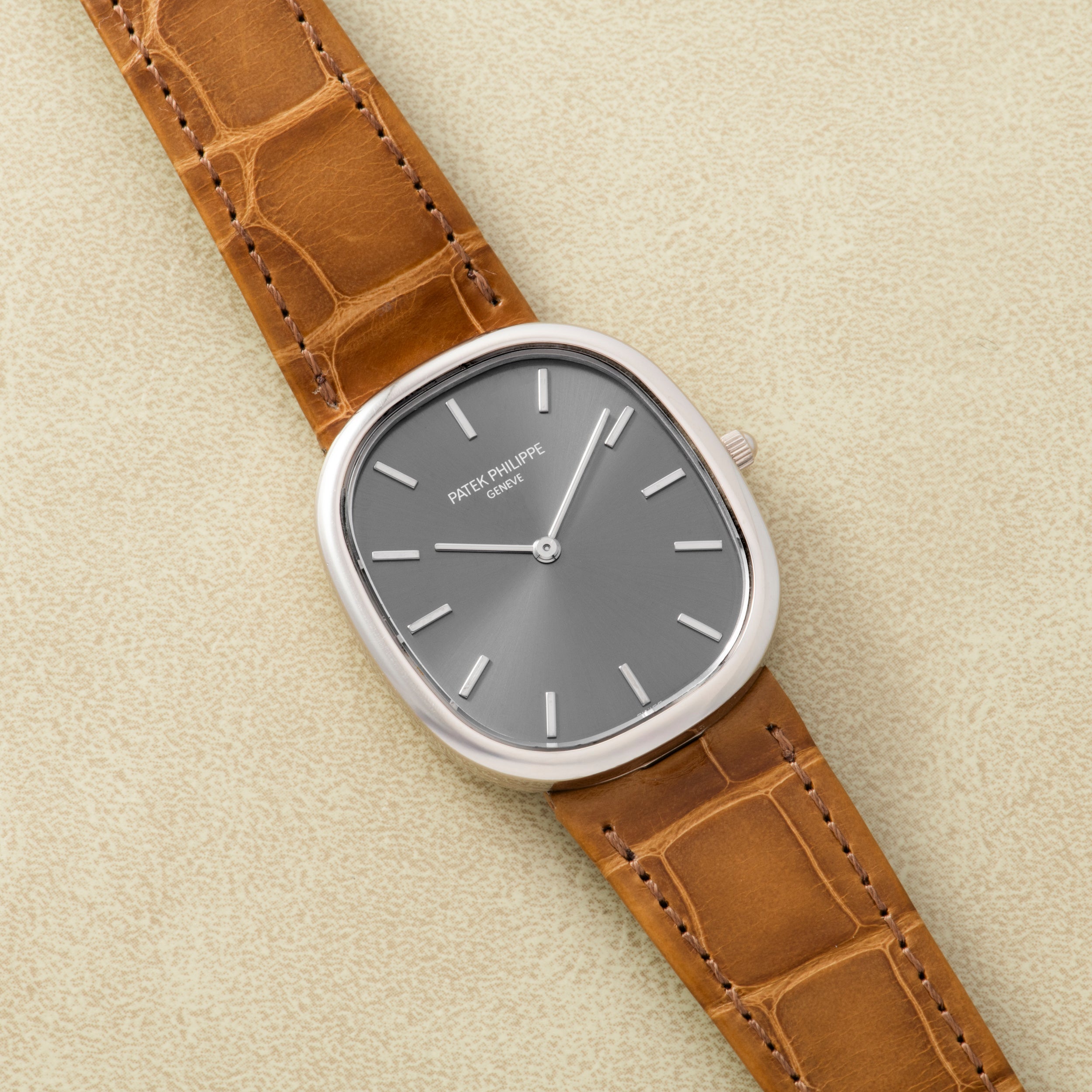 A Patek Philippe Ellipse watch with a silver case, gray dial, and brown leather strap is displayed on a beige surface.
