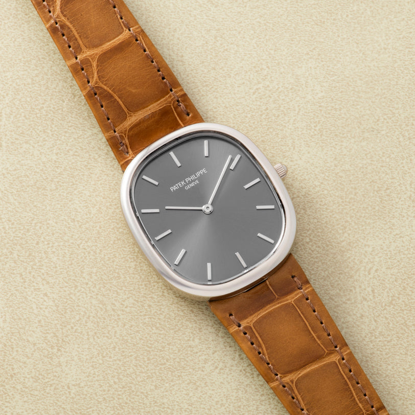 A Patek Philippe Ellipse watch with a silver case, gray dial, and brown leather strap is displayed on a beige surface.