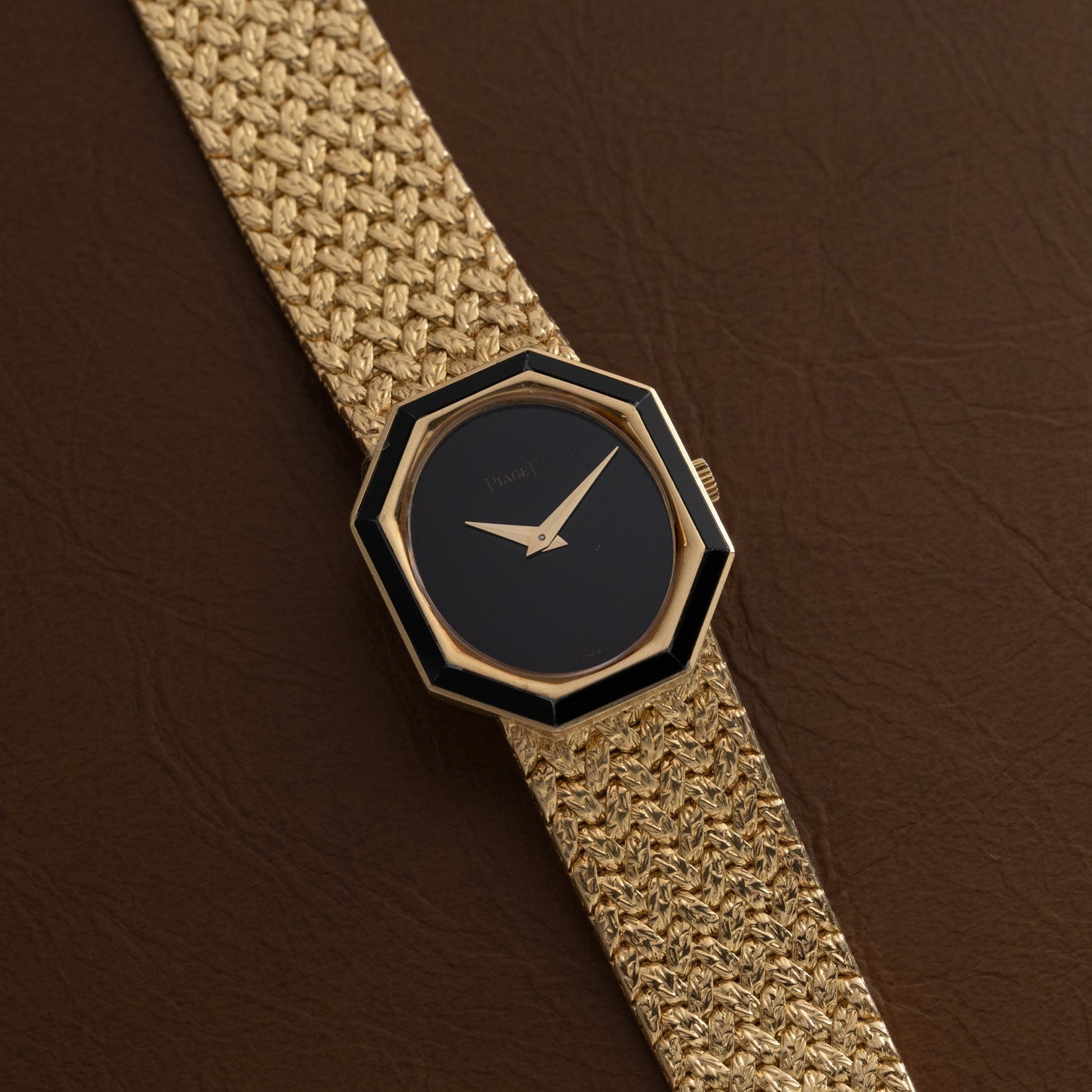 A Piaget Onyx Dial gold watch with a black octagonal face and woven gold band rests on a brown leather surface.