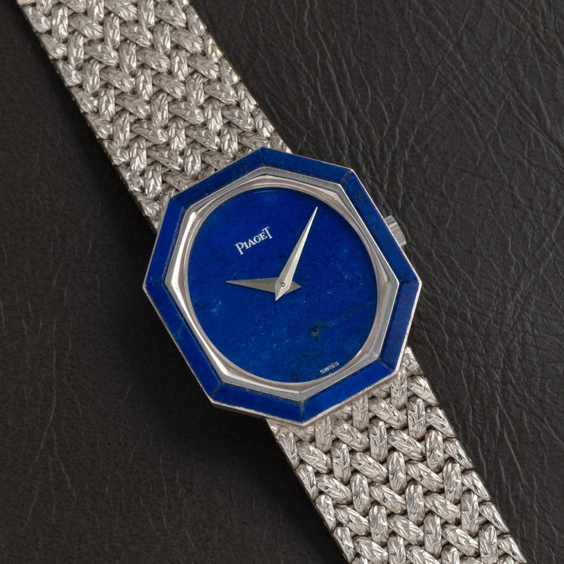 The Piaget Lapis Lazuli White Gold Dress Watch features an octagonal blue face and a woven metal band, displayed on a black leather surface.