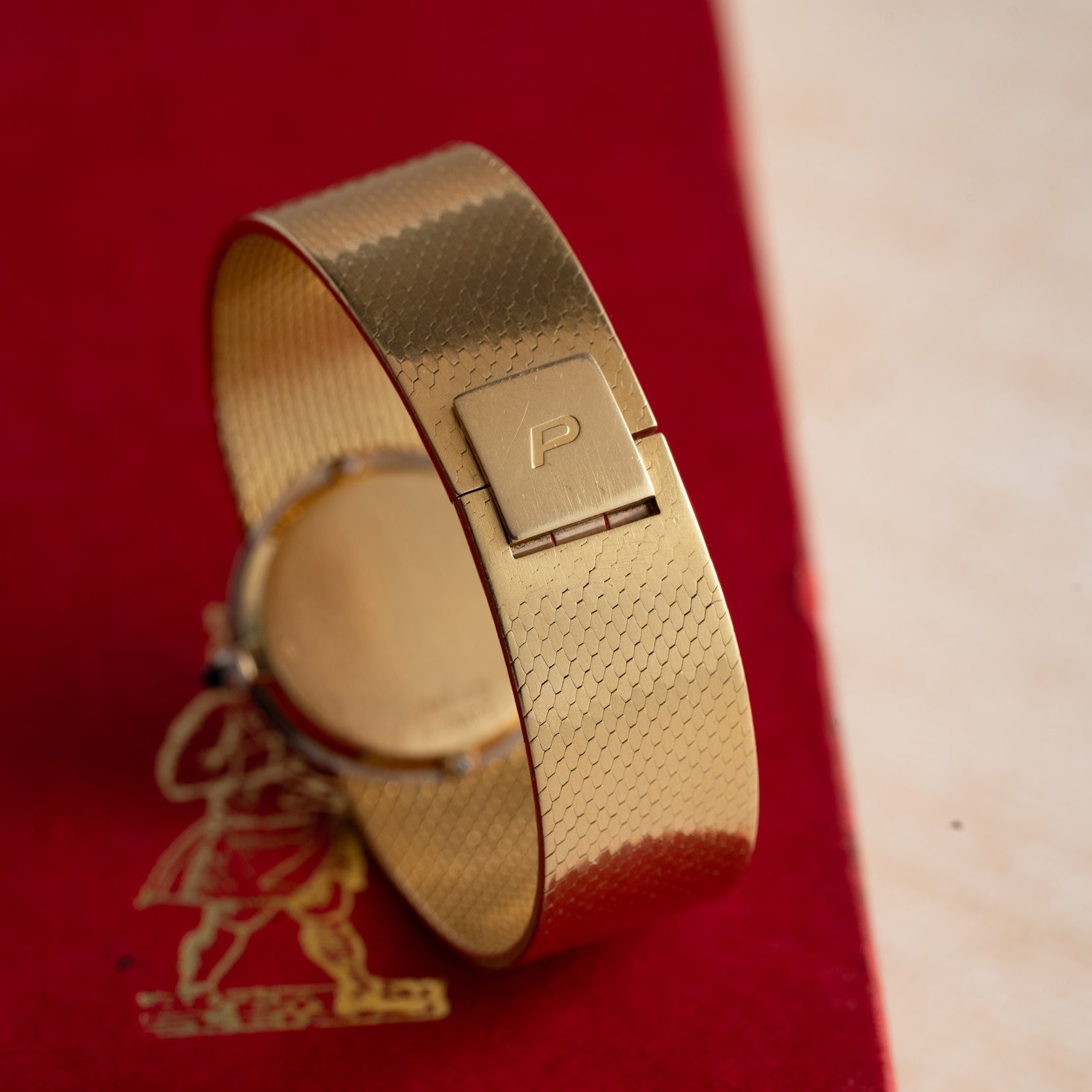 A Piaget Ladies Diamond Watch, Tiffany & Co., featuring a gold textured band and a P-engraved clasp, rests on a red surface.
