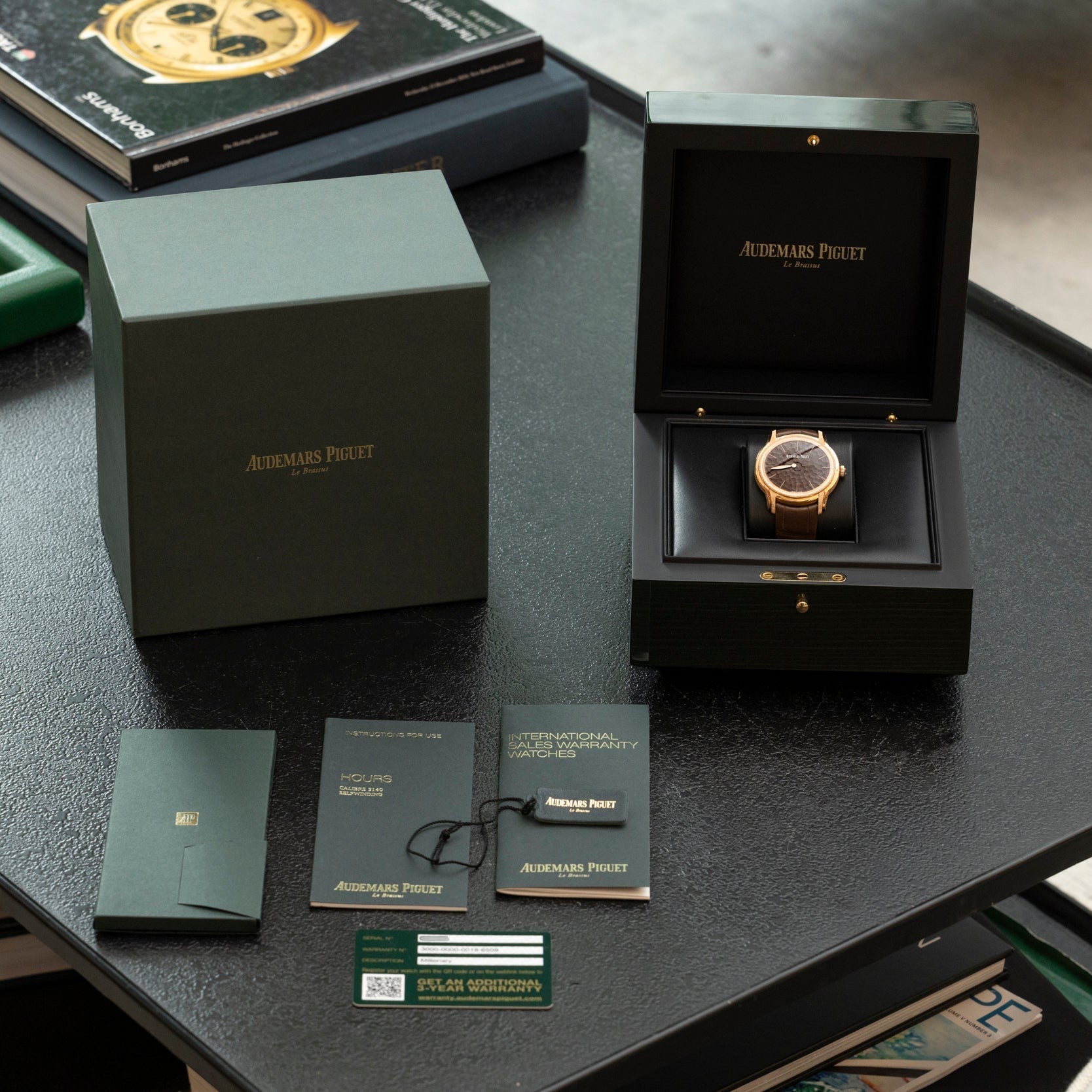 An Audemars Piguet Millenary Philosophique watch is shown in its box, with original packaging, manuals, and certificates displayed on a table.