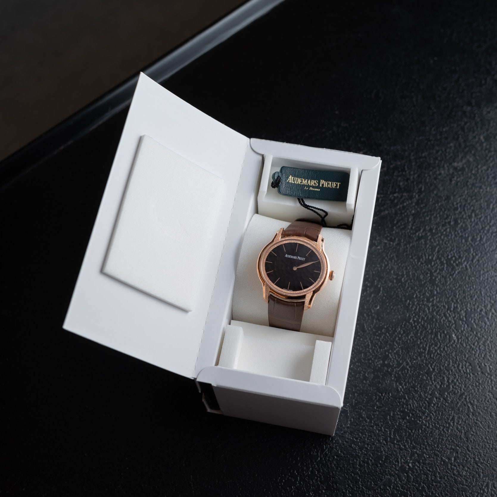 An Audemars Piguet Millenary Philosophique wristwatch in a brown tone is displayed in an open white box on a black surface.