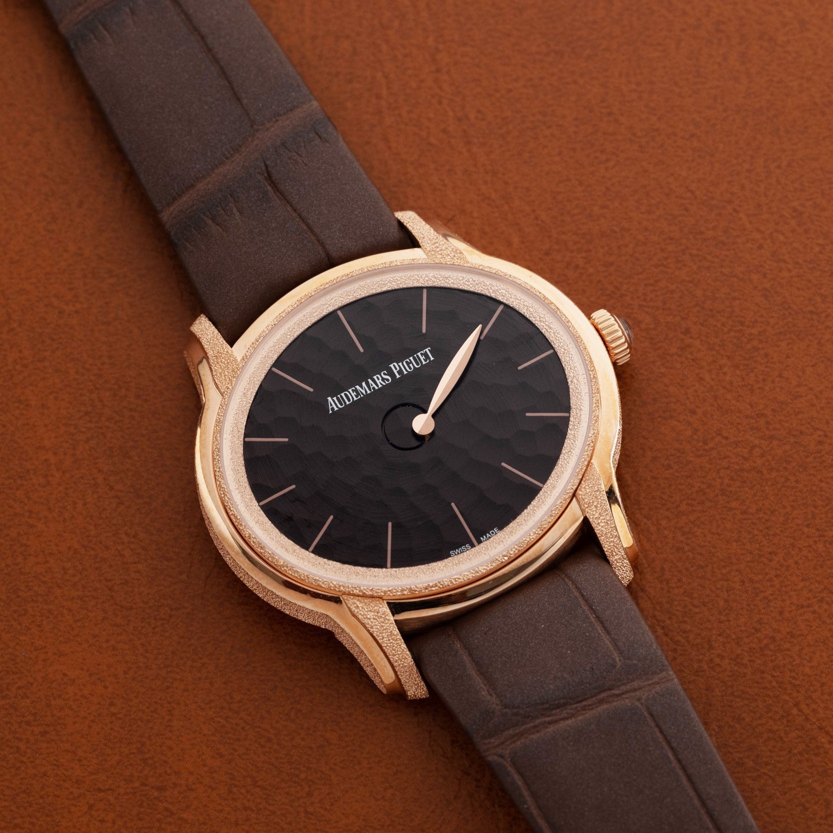 The Audemars Piguet Millenary Philosophique features a gold case, black textured dial, and brown leather strap, displayed against a brown background.