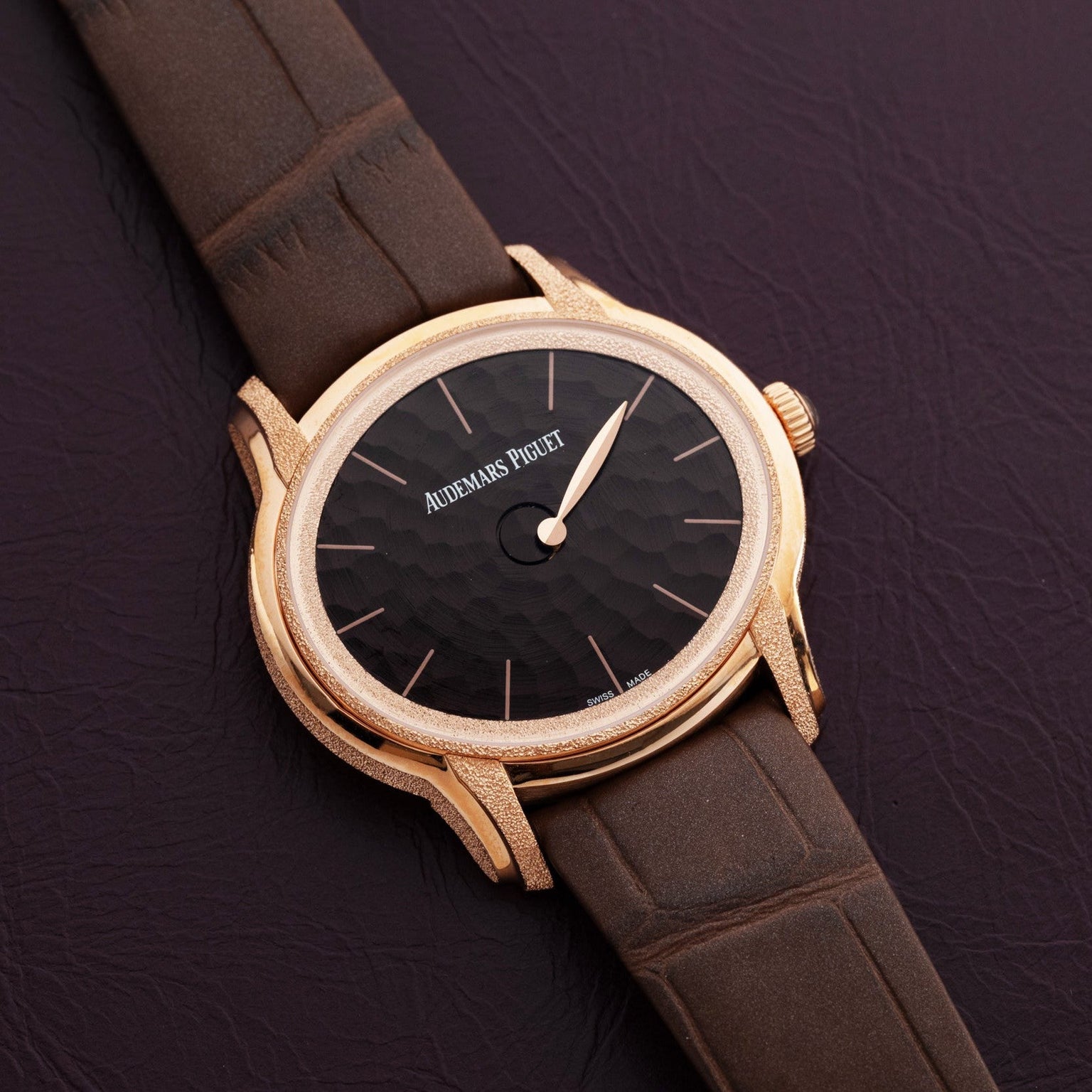 The Audemars Piguet Millenary Philosophique, featuring a gold case, black textured dial, and brown leather strap, is displayed on a dark leather surface.
