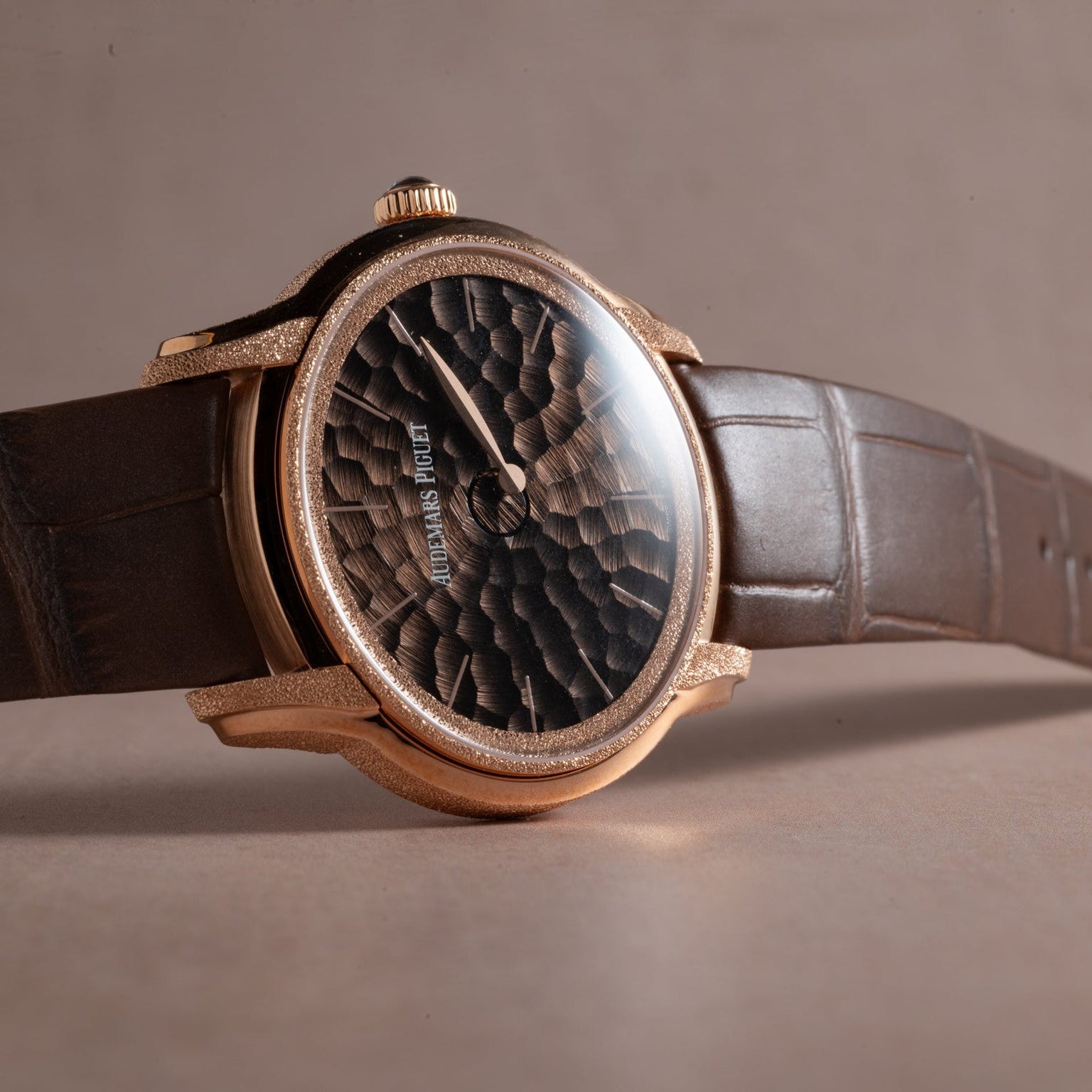 The Audemars Piguet Millenary Philosophique features a textured black dial, gold case, and brown leather strap, displayed on a beige surface.