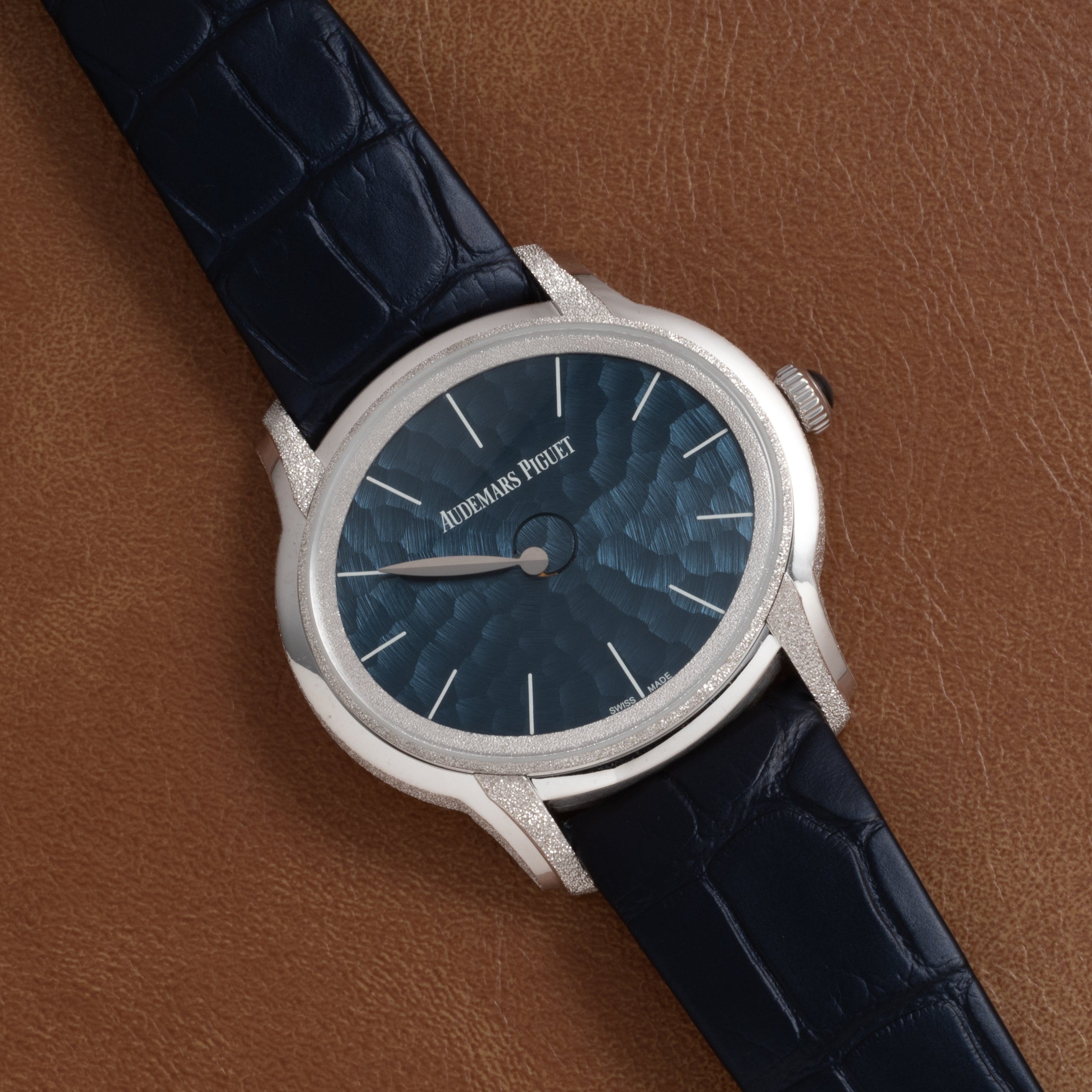 The Audemars Piguet Millenary Philosophique features a blue textured dial and black leather strap, displayed on a brown leather background.