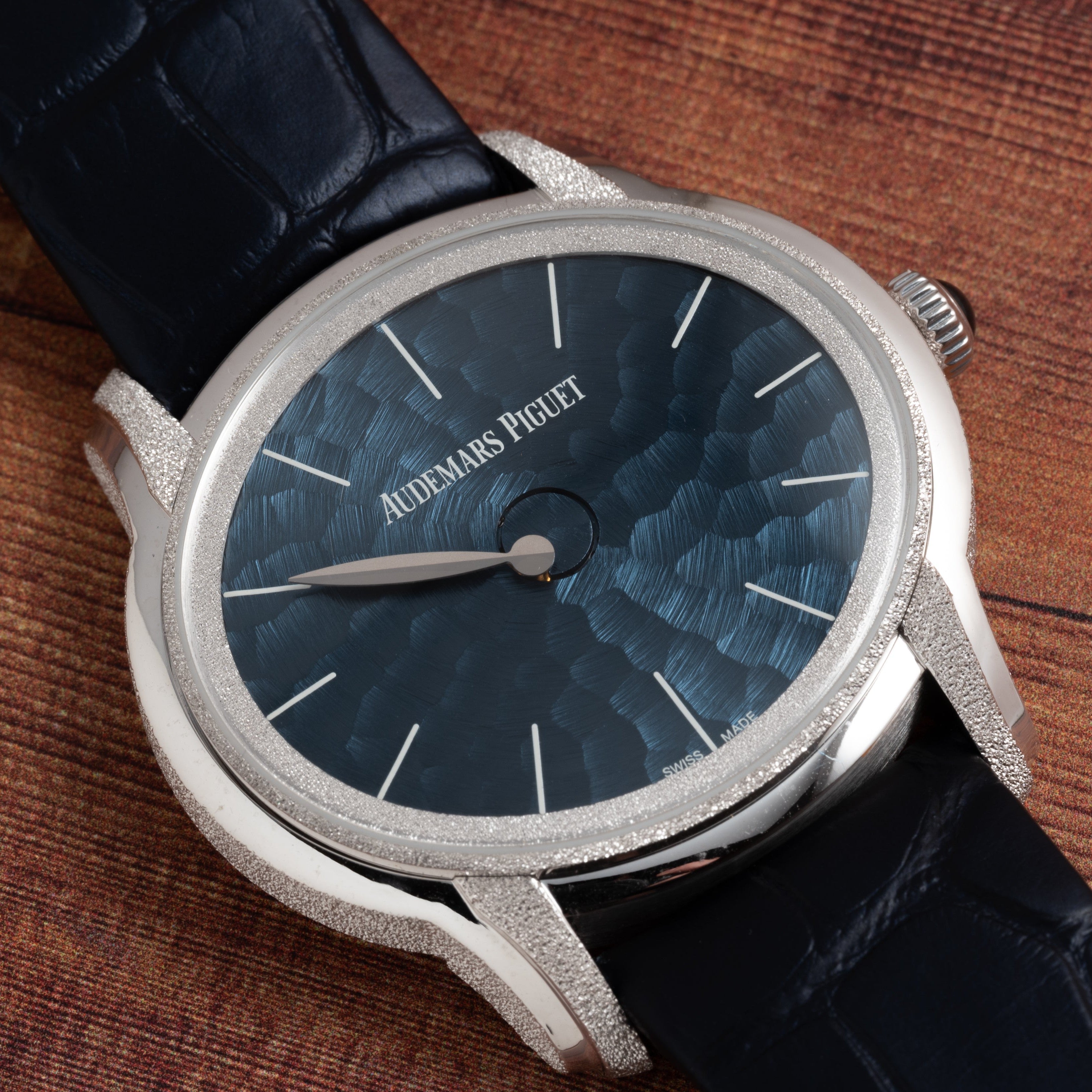 The Audemars Piguet Millenary Philosophique watch features a textured blue dial and a black leather strap, displayed on a wooden surface.