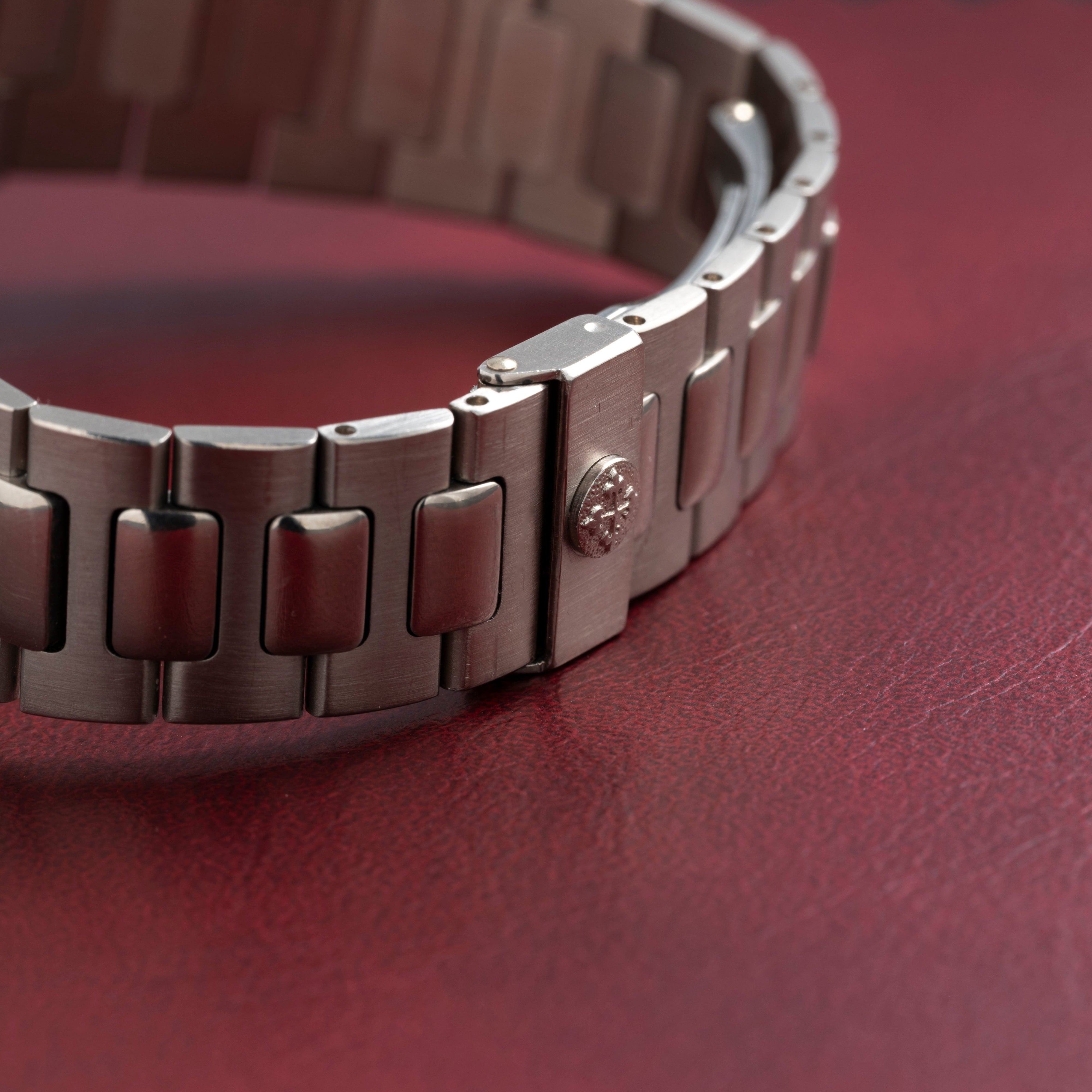 Close-up of a metal watch bracelet with a Patek Philippe Nautilus logo on a textured red surface.