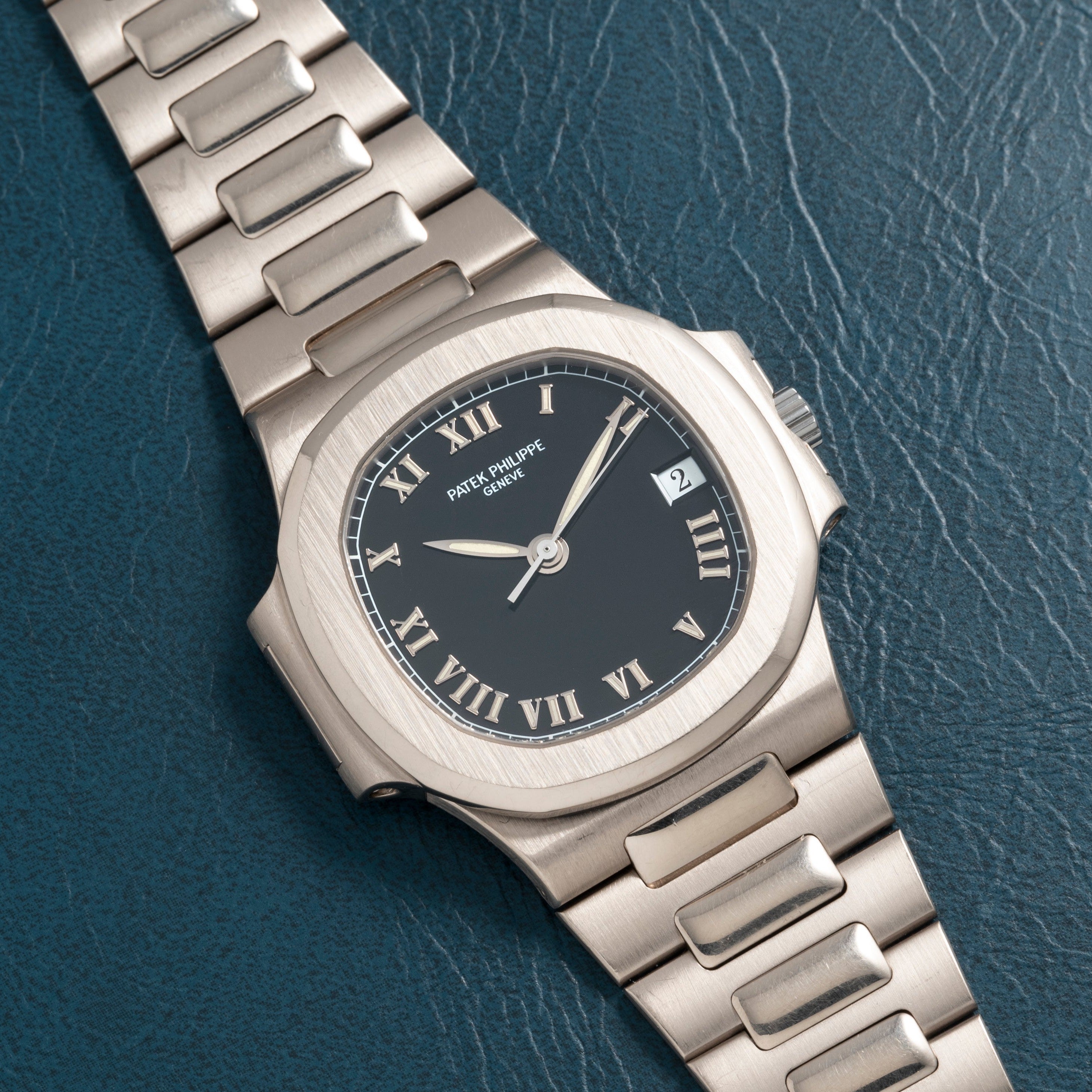 A silver Patek Philippe Nautilus wristwatch with Roman numerals on a dark face, resting on textured dark leather.