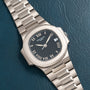 A silver Patek Philippe Nautilus wristwatch with Roman numerals on a dark face, resting on textured dark leather.