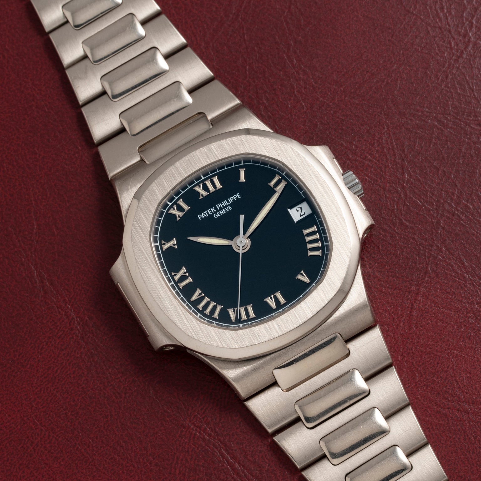 A Patek Philippe Nautilus silver wristwatch with a black face and Roman numerals placed on a red textured surface.