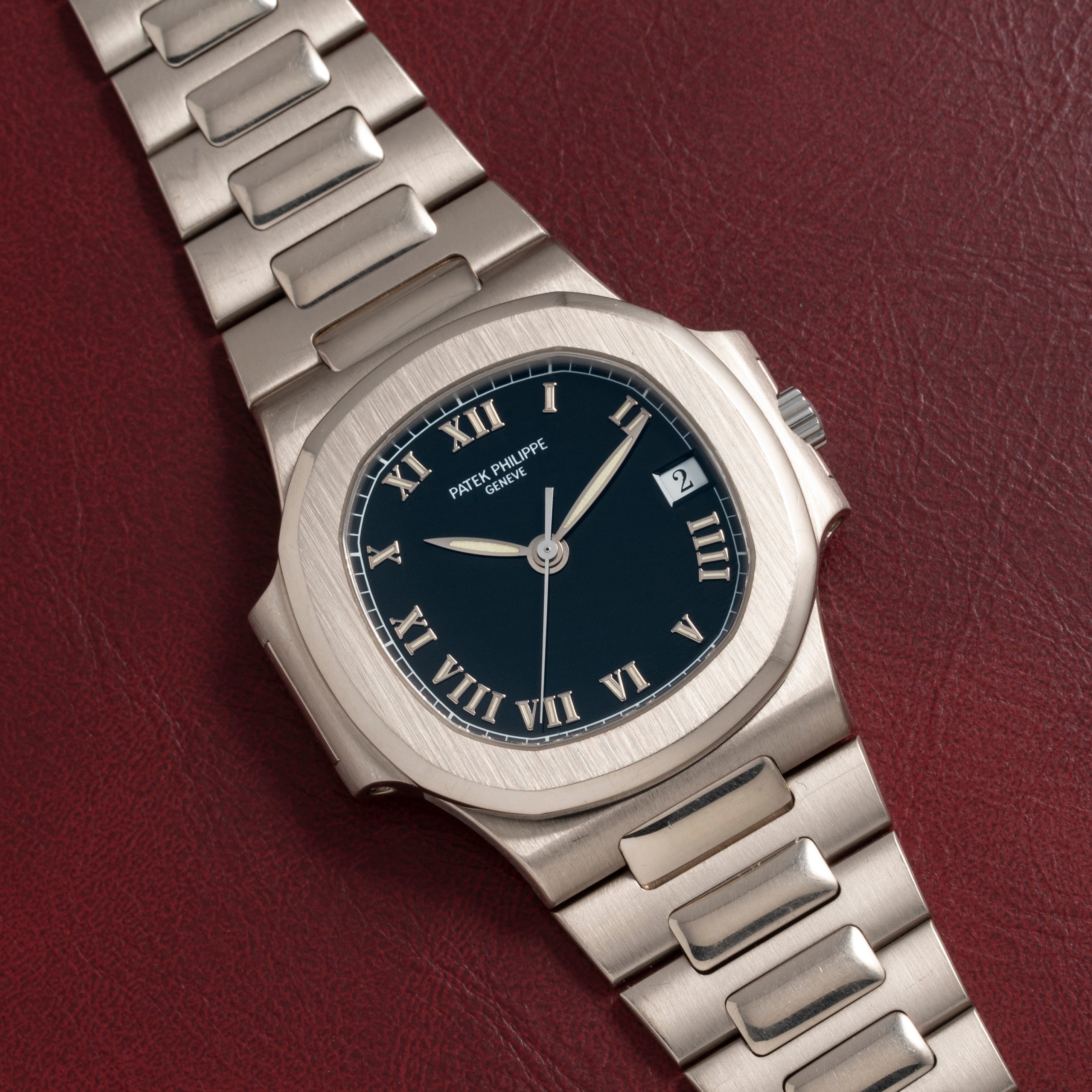 A Patek Philippe Nautilus silver wristwatch with a black face and Roman numerals placed on a red textured surface.
