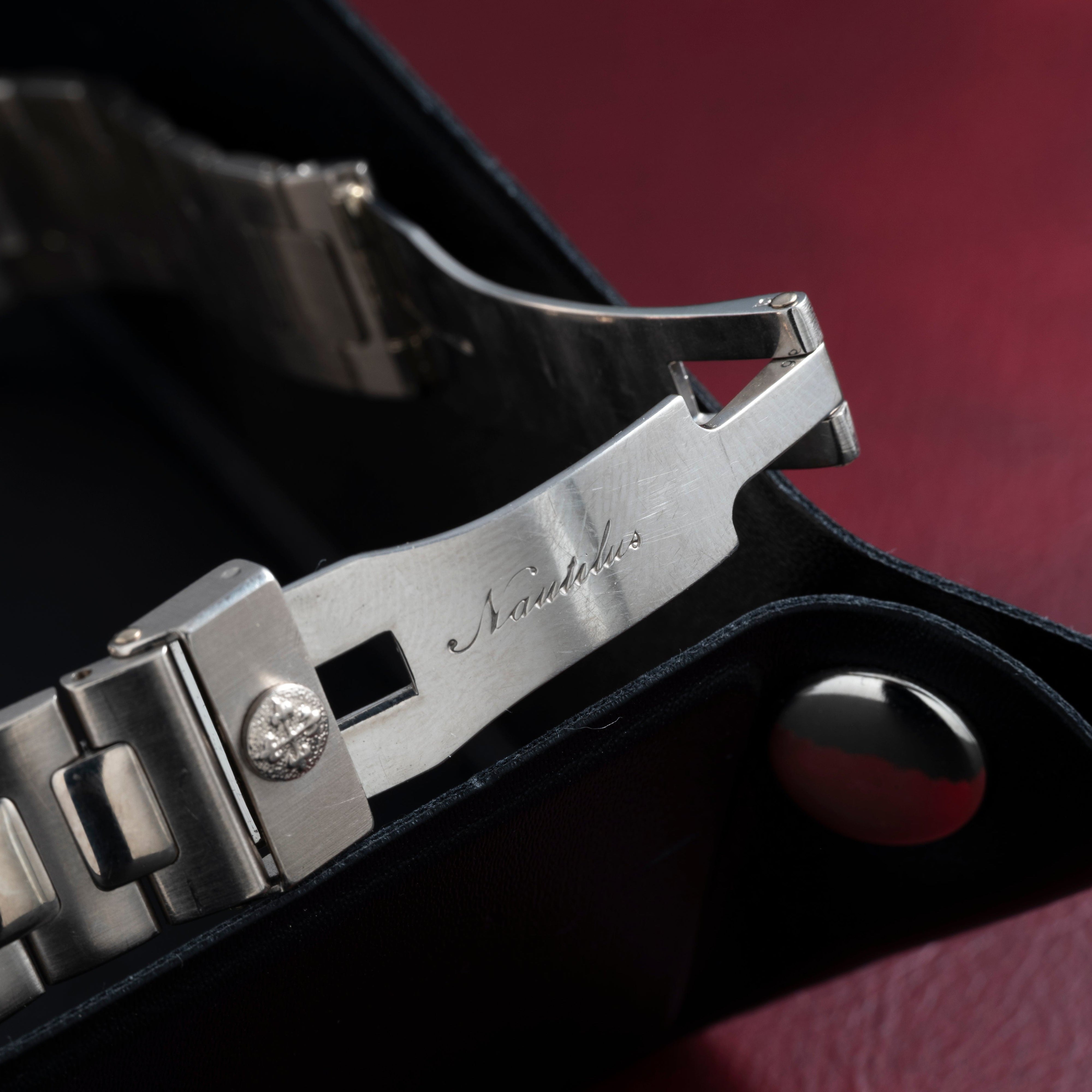 Close-up of a metal Patek Philippe Nautilus watch clasp engraved with Nautilus against a red and black background.
