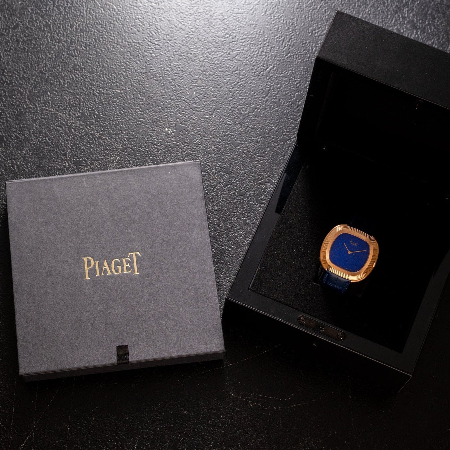 A Piaget Black Tie Andy Warhol gold and blue watch in a black box sits beside a closed Piaget case on a dark surface.