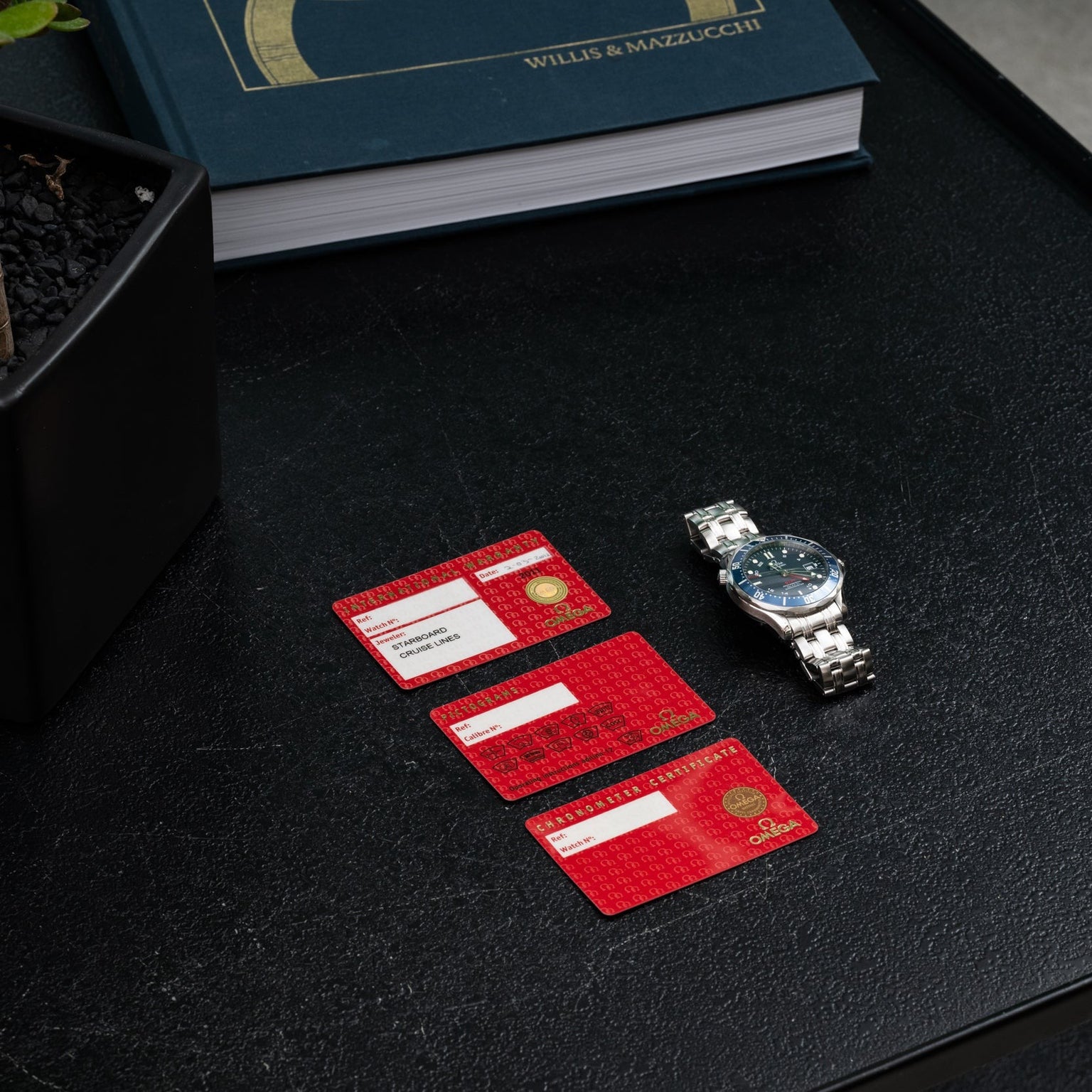 Three red cards and an OMEGA Seamaster 300 Professional GMT wristwatch rest on a black table beside a book and a plant.