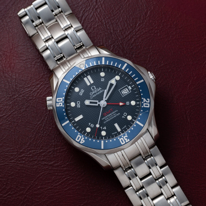 An OMEGA Seamaster 300 Professional GMT watch with a blue bezel rests on a maroon leather surface.