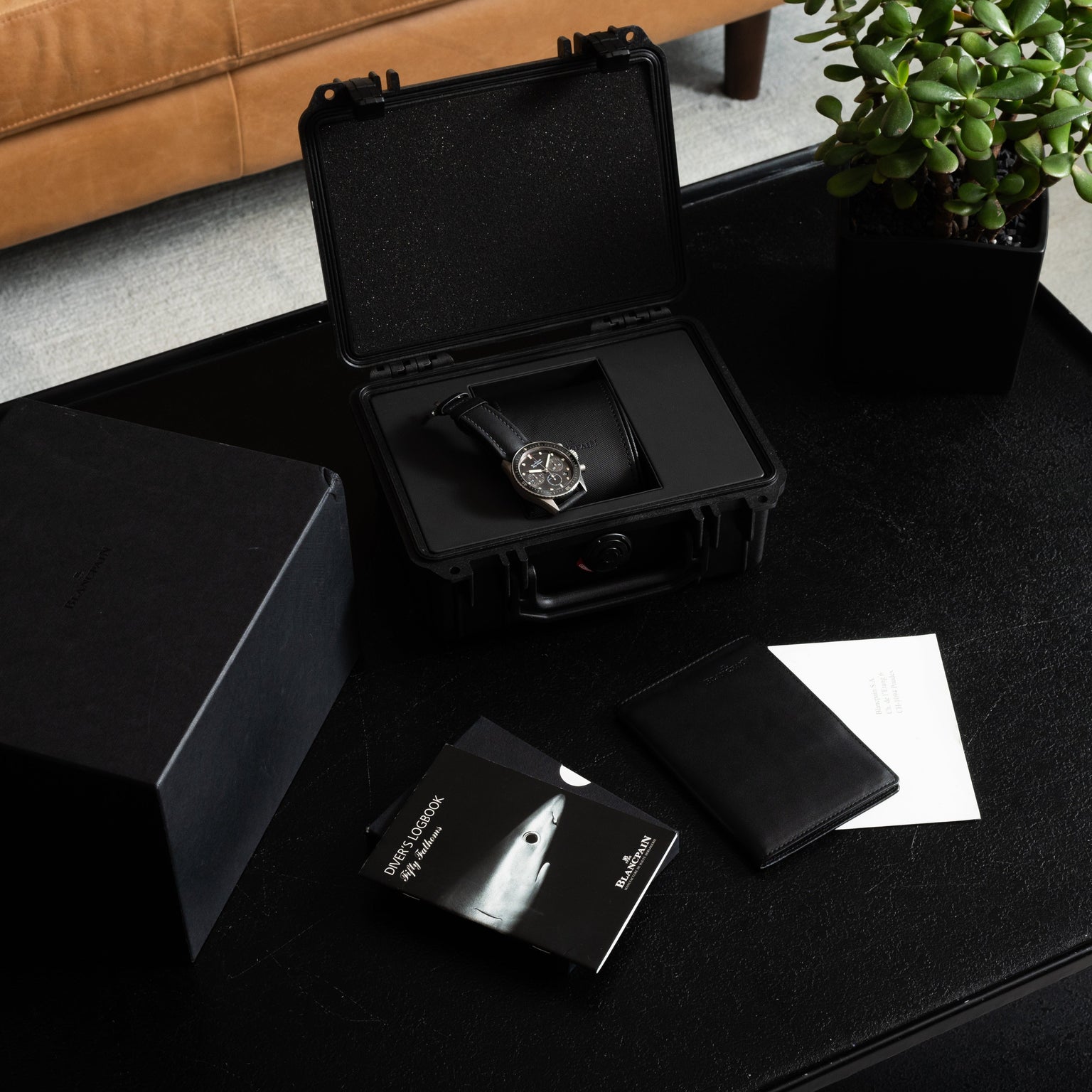 A Blancpain Fifty Fathoms Bathyscaphe Chronographe Flyback wristwatch in a black case rests on a table, accompanied by booklets, a wallet, and a nearby potted plant.