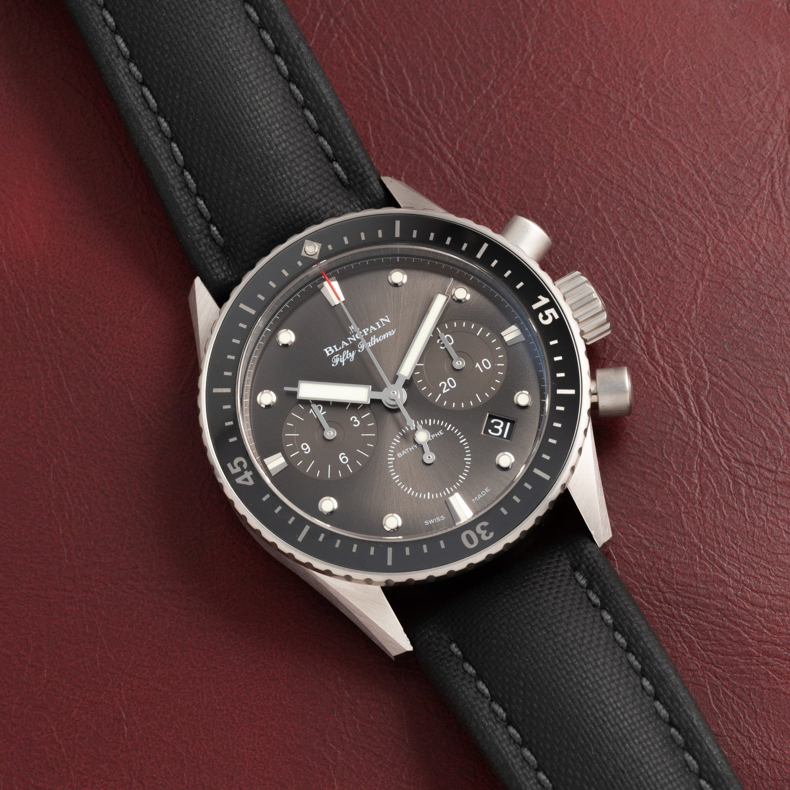 The Blancpain Fifty Fathoms Bathyscaphe Chronographe Flyback, a black chronograph wristwatch with a matching strap, is displayed on a dark red leather surface.