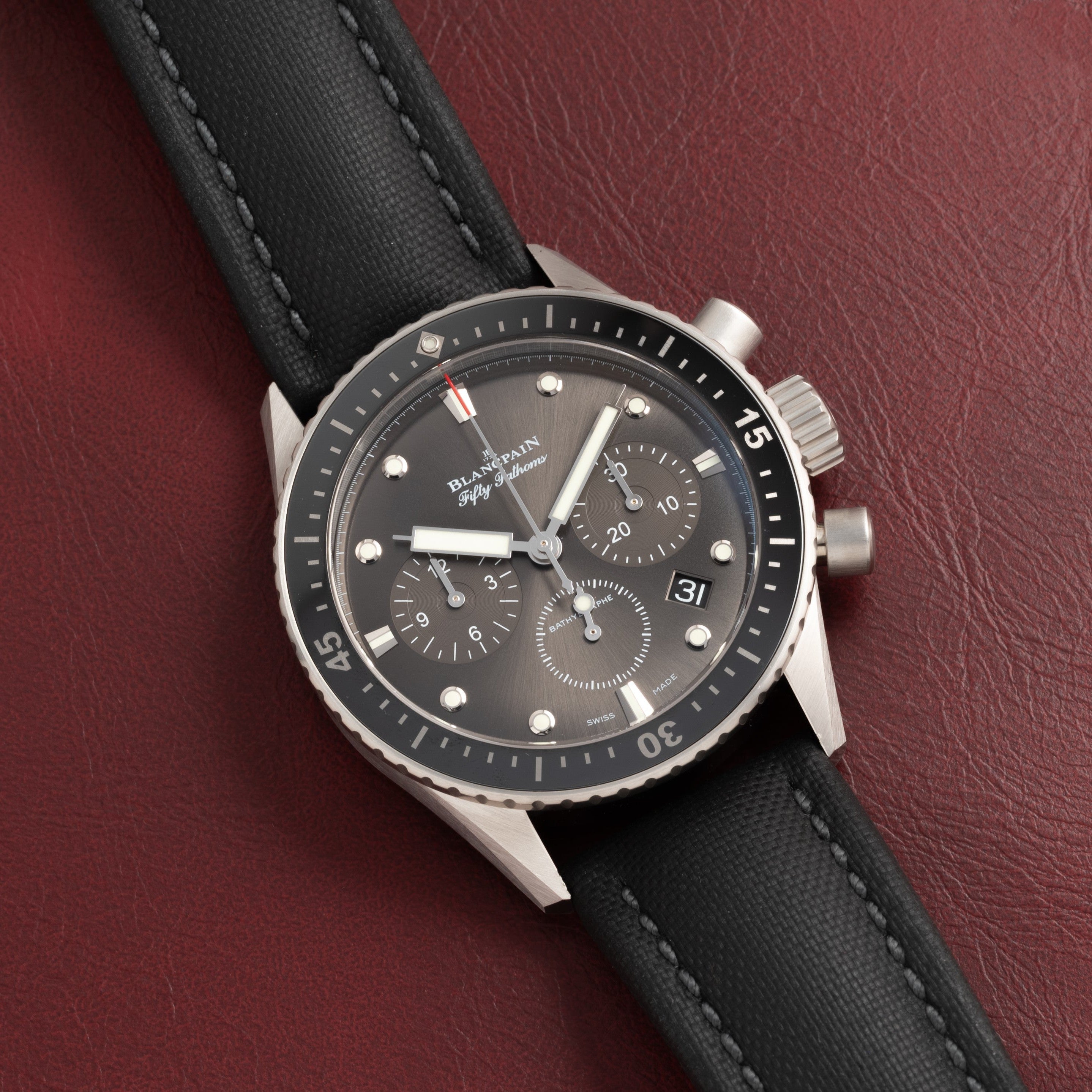 The Blancpain Fifty Fathoms Bathyscaphe Chronographe Flyback, a black chronograph wristwatch with a matching strap, is displayed on a dark red leather surface.