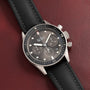 The Blancpain Fifty Fathoms Bathyscaphe Chronographe Flyback, a black chronograph wristwatch with a matching strap, is displayed on a dark red leather surface.