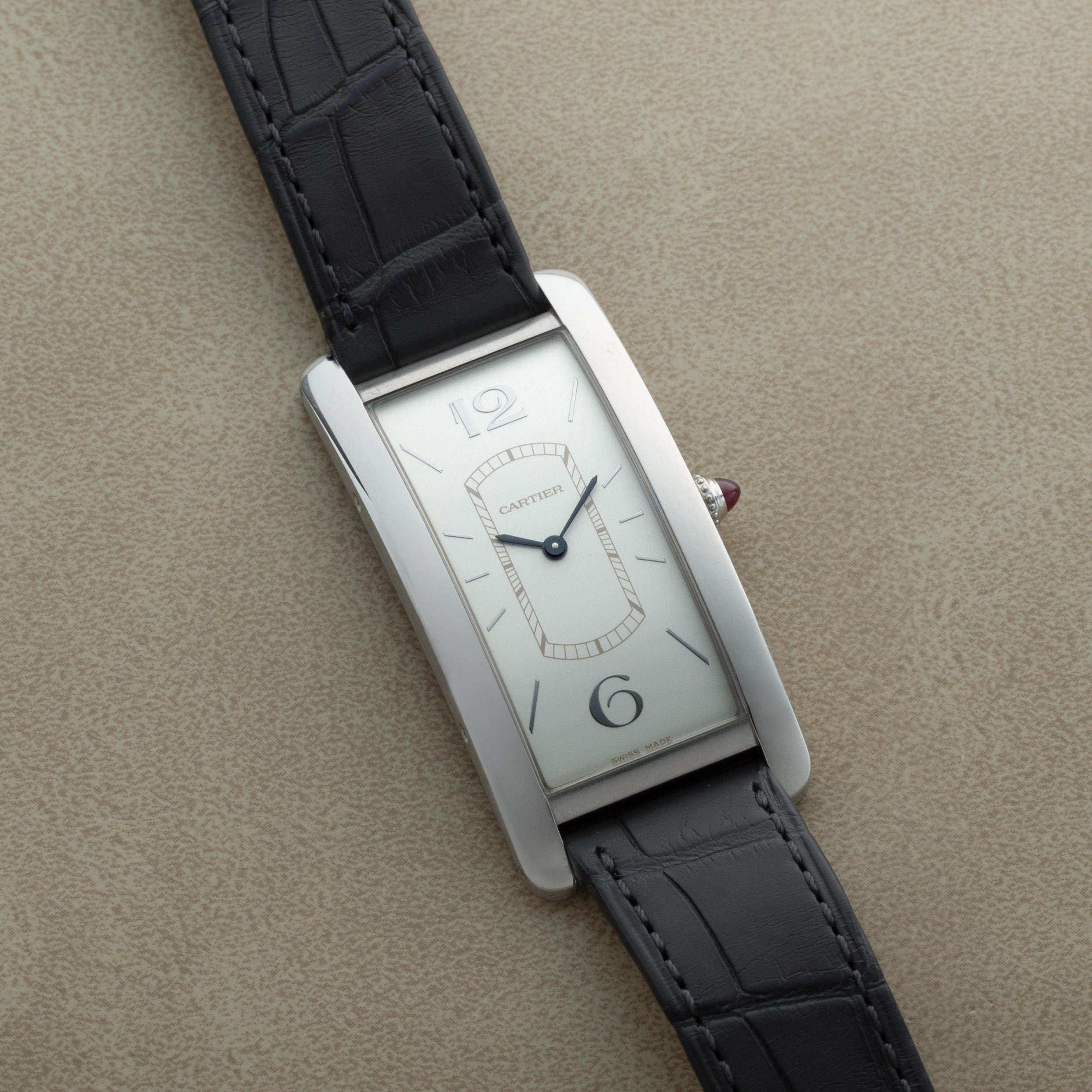 A Cartier Tank Cintrée Limited Edition rectangular wristwatch with a silver face and black leather strap displayed on a beige surface.