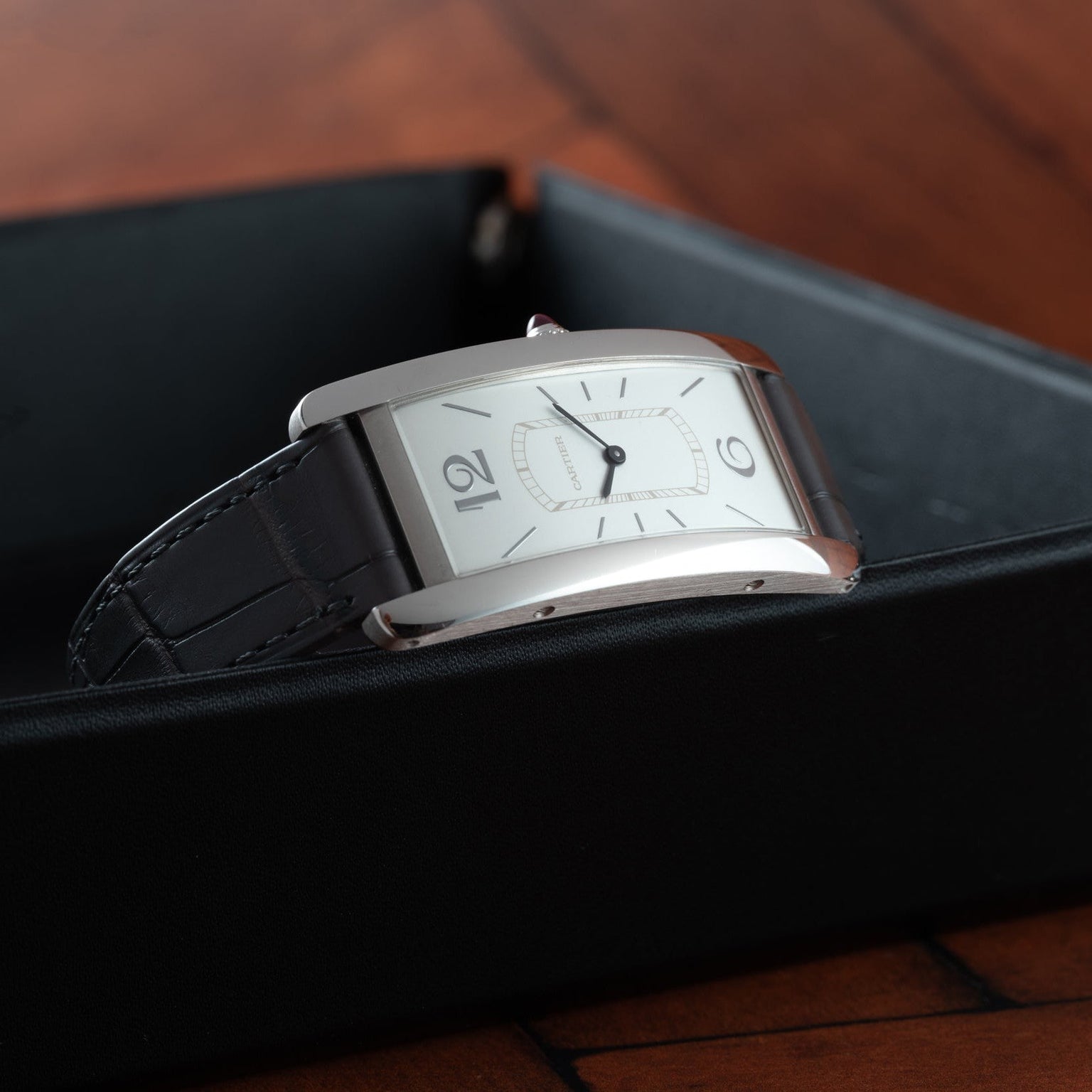 The Cartier Tank Cintrée Limited Edition, a rectangular silver wristwatch with a black leather strap, rests on a black box.