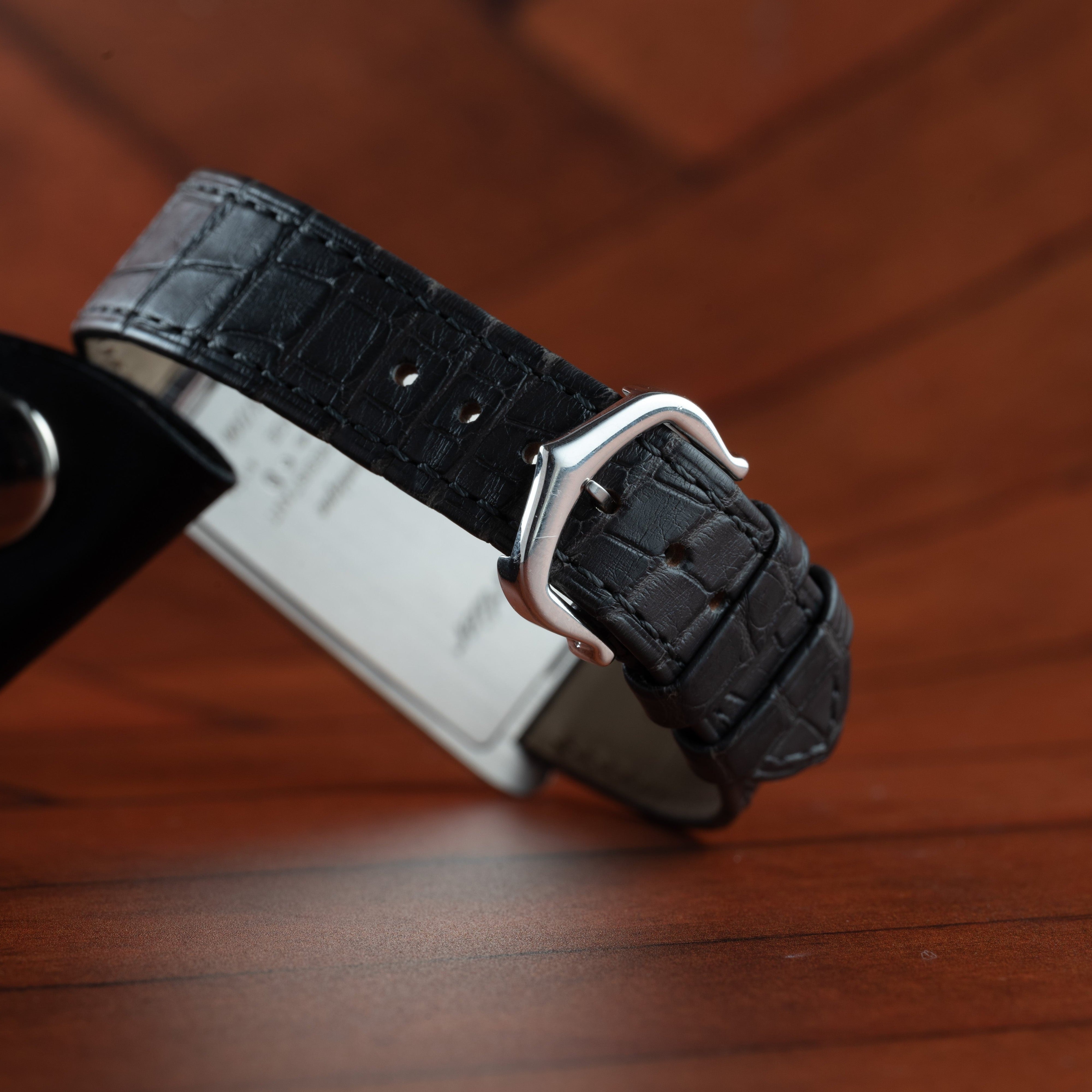 The Cartier Tank Cintrée Limited Edition black leather watch strap with a silver buckle is displayed on a wooden surface.