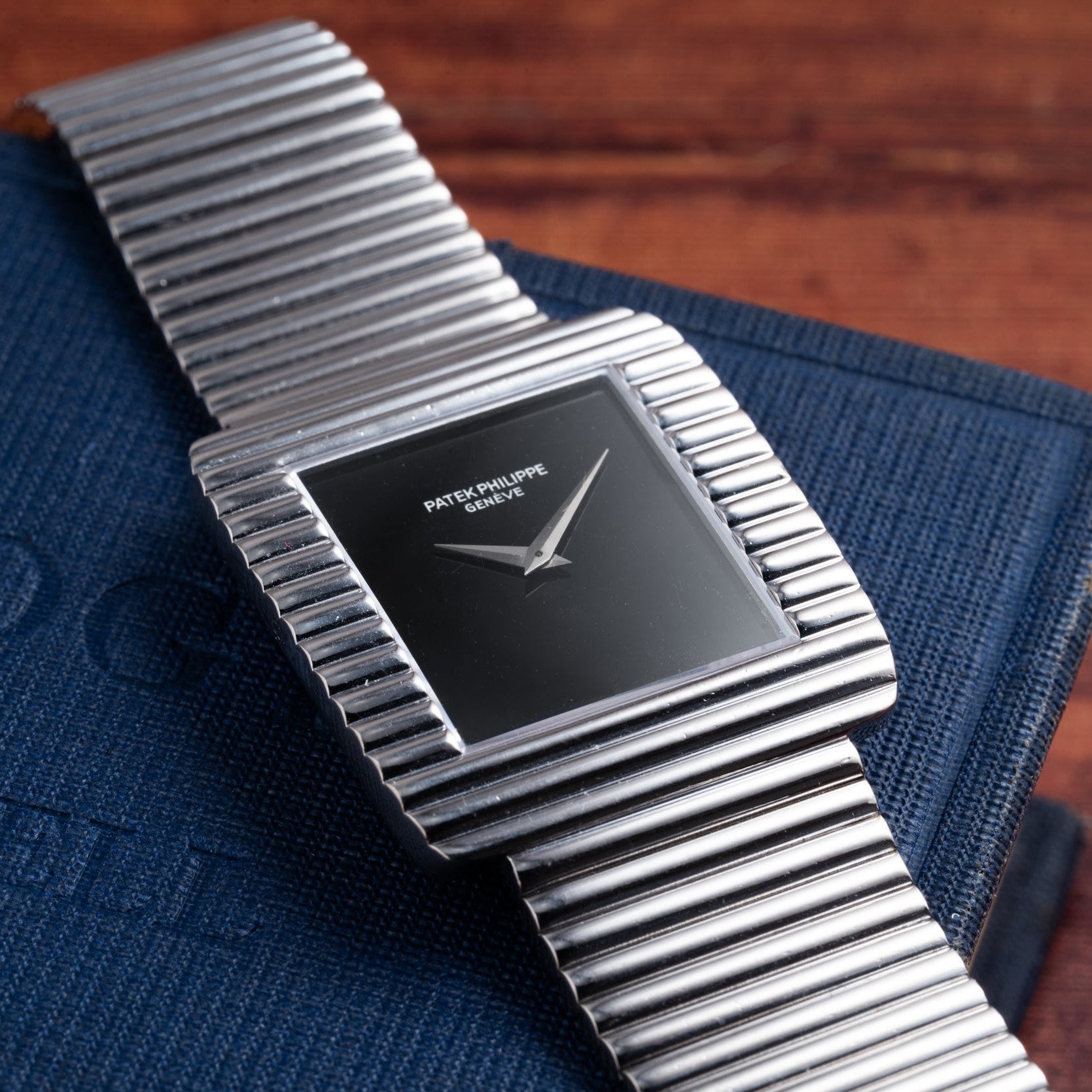 A Patek Philippe Ellipse Gondolo Onyx watch with a square silver case and black dial on a ribbed metal band, displayed on a blue book.