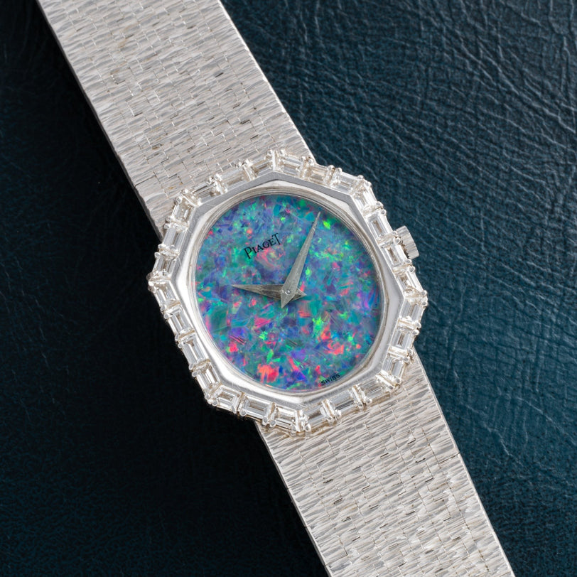 The Piaget Opal White Gold Dress Watch features a colorful opal dial and gem-set bezel, set on a textured band, resting on dark leather.