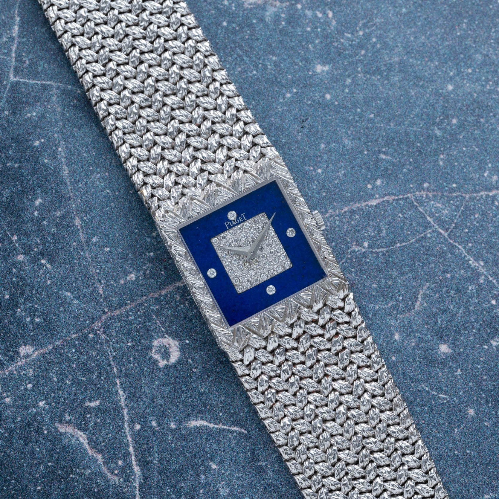 The Piaget Tradition Lapis Lazuli Diamond Dress Watch features a blue square face and textured silver band, exuding luxury on a marble surface.