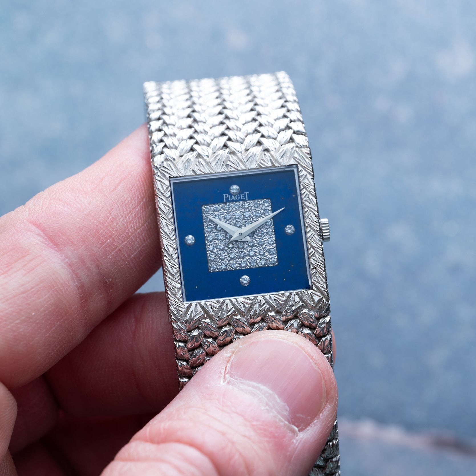 A hand holding a Piaget Tradition Lapis Lazuli Diamond Dress Watch with a blue face and diamond-studded square center.