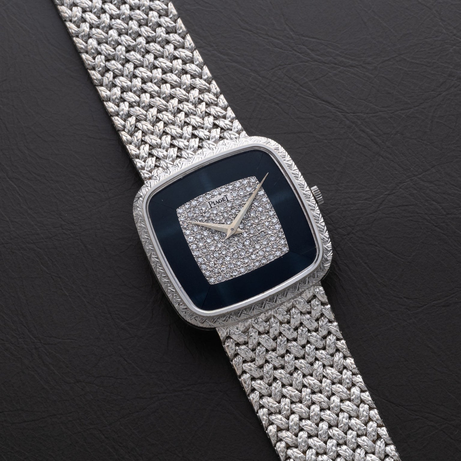 A Piaget Hawk Eye White Gold Dress Watch with a diamond-studded face and woven metal band displayed on a black surface.