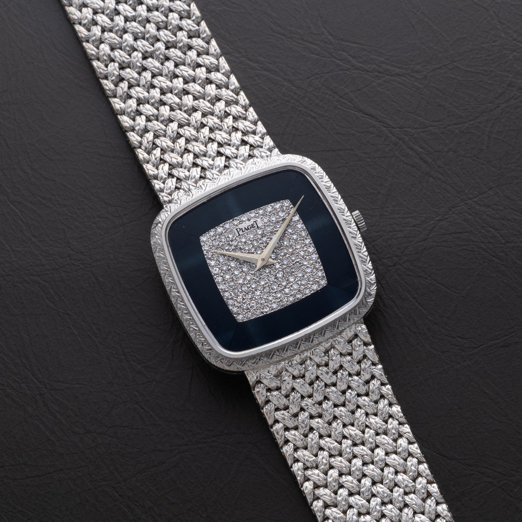 A Piaget Hawk Eye White Gold Dress Watch with a diamond-studded face and woven metal band displayed on a black surface.