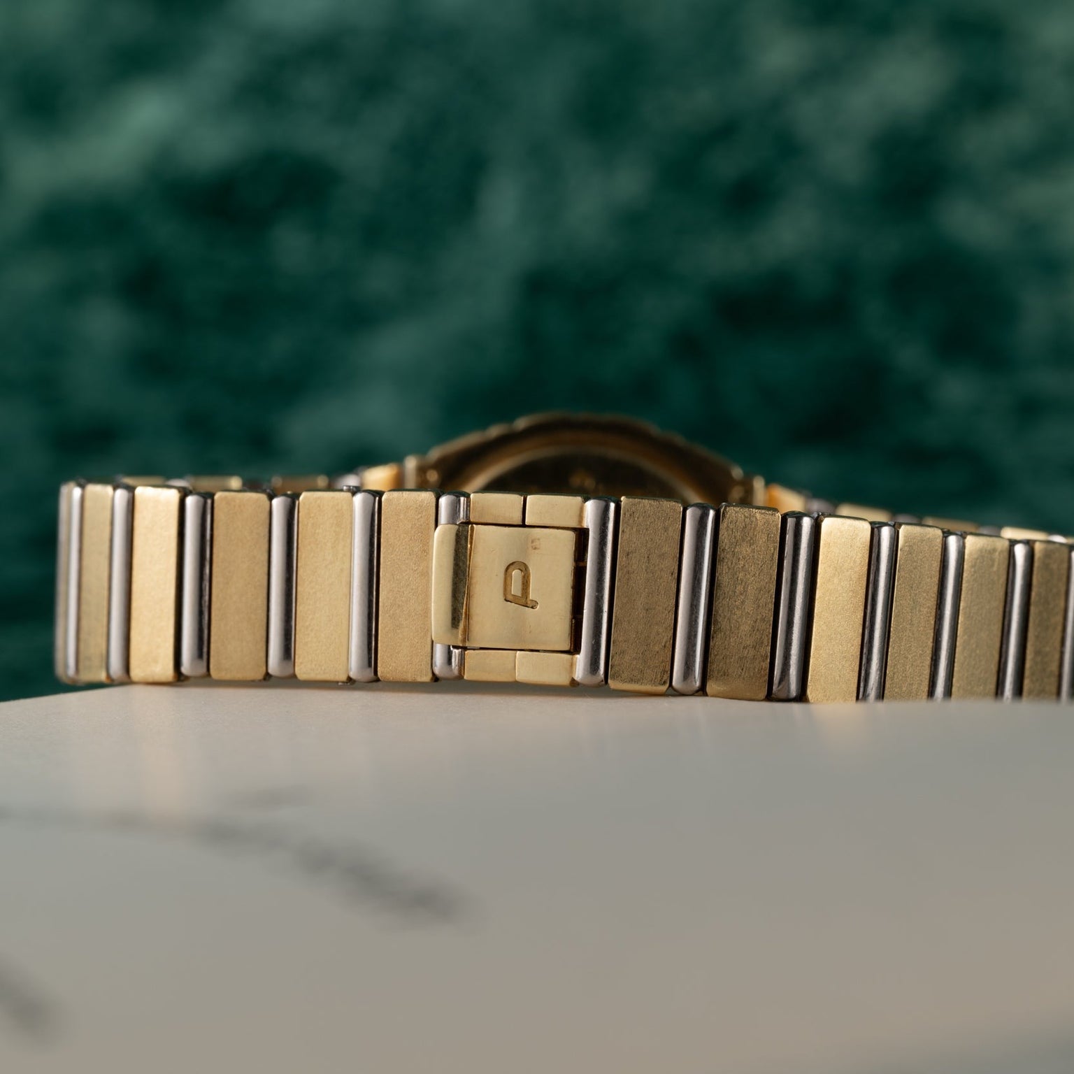 Close-up of the Piaget Polo Two-Tone watch band, featuring a P engraved on the clasp, set against a green background.