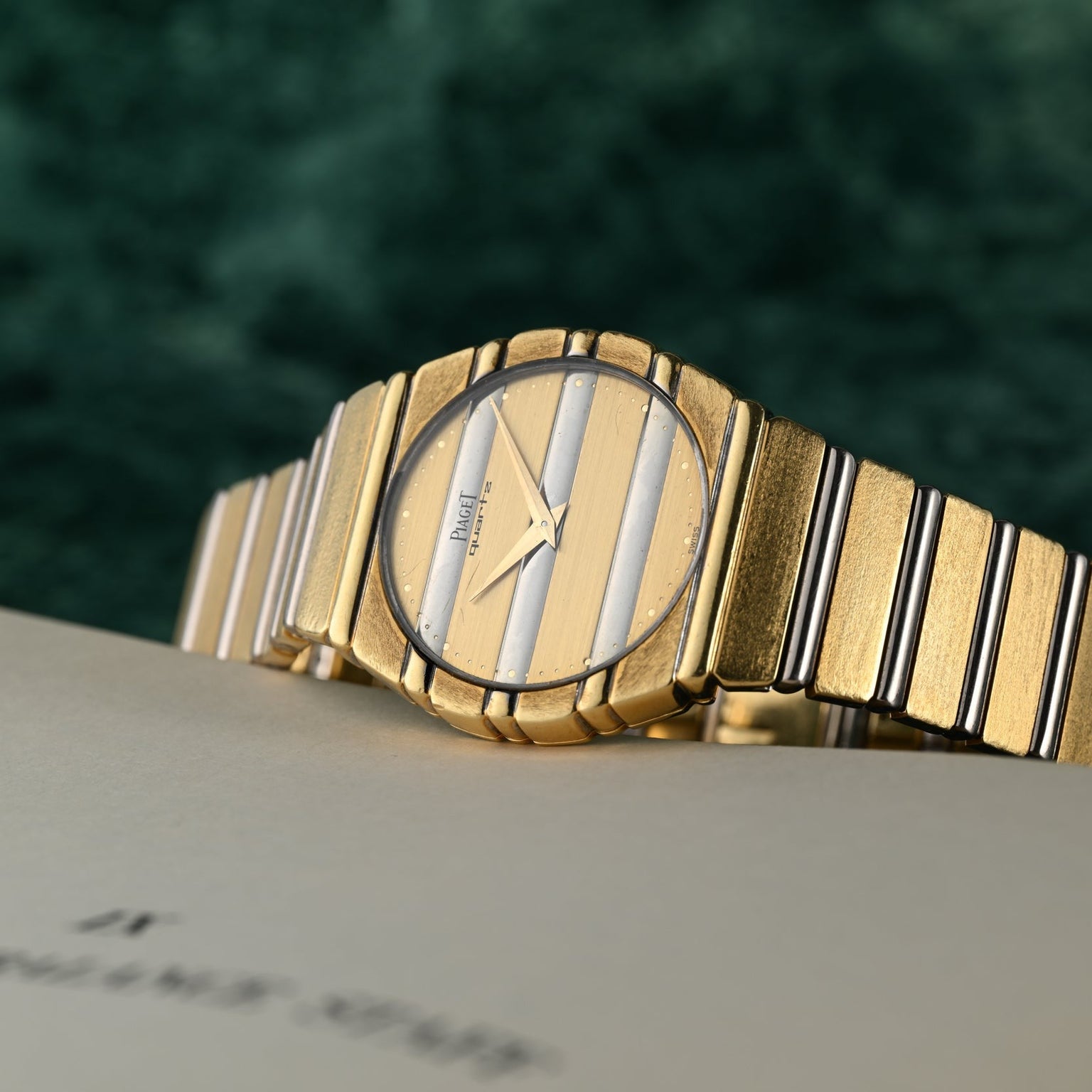 The Piaget Polo Two-Tone luxury wristwatch, featuring gold and silver stripes, is displayed on a cream-colored surface with a green background.