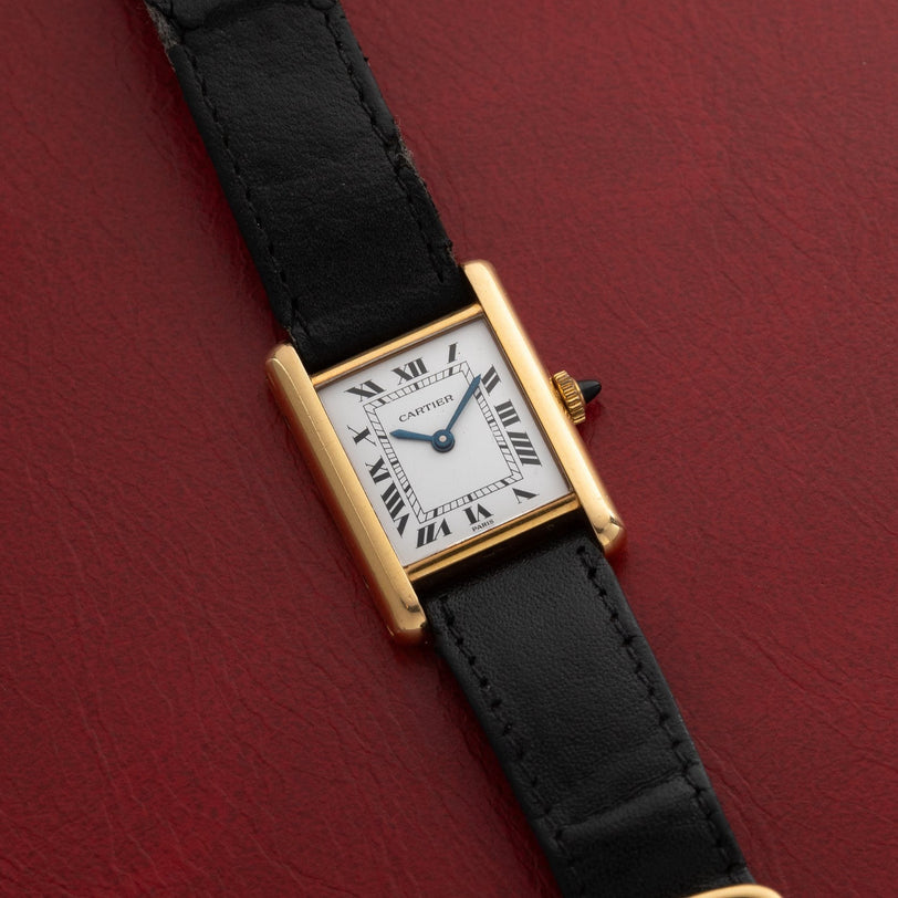 Cartier Tank Piccolo watch featuring a white dial with black Roman numerals and a black leather strap, displayed on a red surface.
