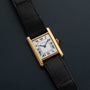 A Cartier Tank Piccolo gold watch with a black leather strap and Roman numeral dial rests on a dark surface.
