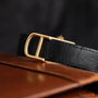A close-up of the Cartier Tank Piccolo black leather belt with a gold buckle, resting on a brown leather surface.