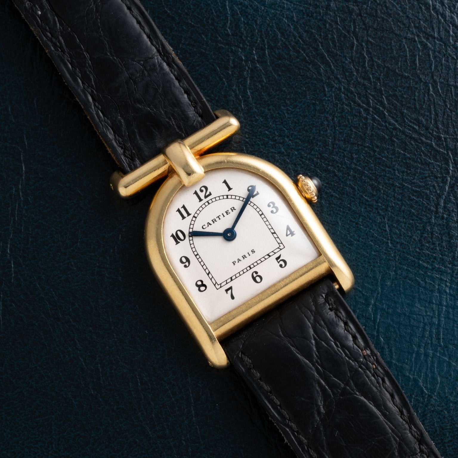 A Cartier Calandre gold wristwatch with a black leather strap rests on a dark textured surface.