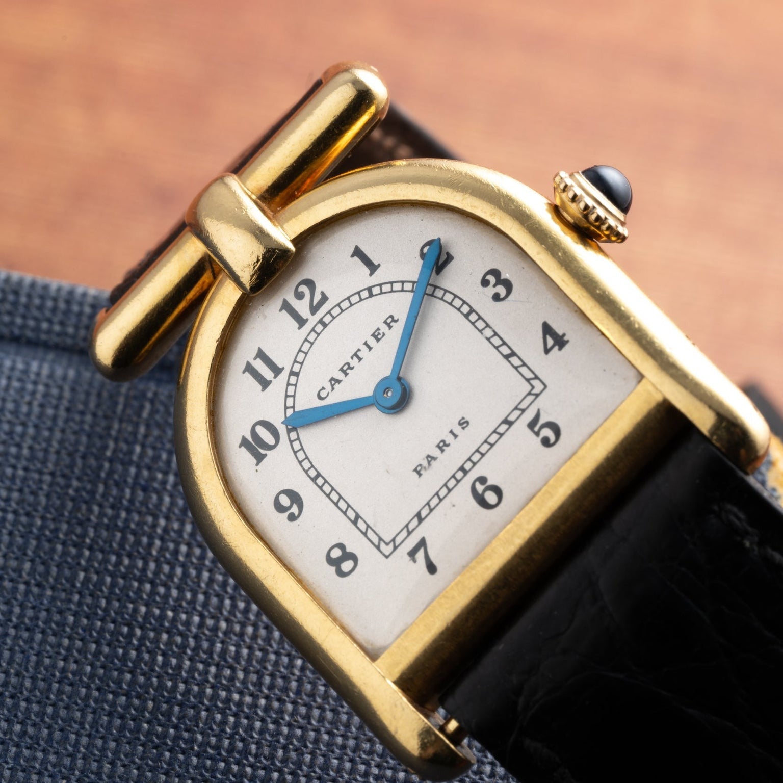 Cartier Calandre gold wristwatch featuring a white dial, black strap, and blue hand, displayed on a blue fabric surface.