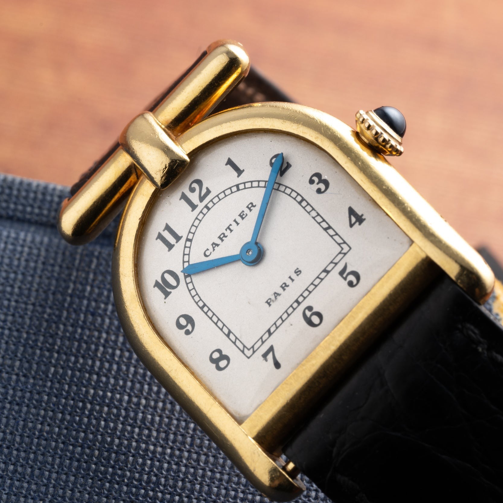 Cartier Calandre gold wristwatch featuring a white dial, black strap, and blue hand, displayed on a blue fabric surface.