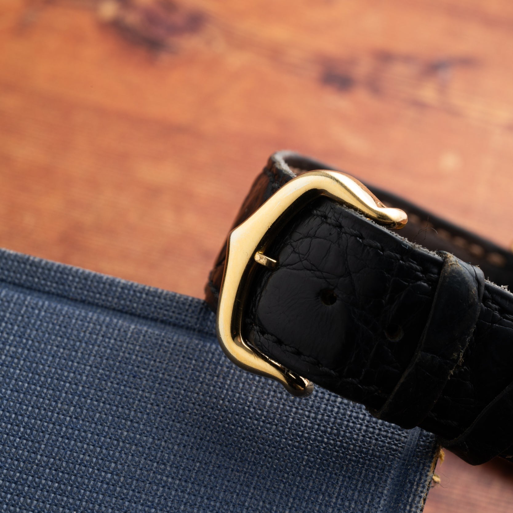The Cartier Calandre black leather watch strap with a gold buckle is displayed on a blue fabric surface.