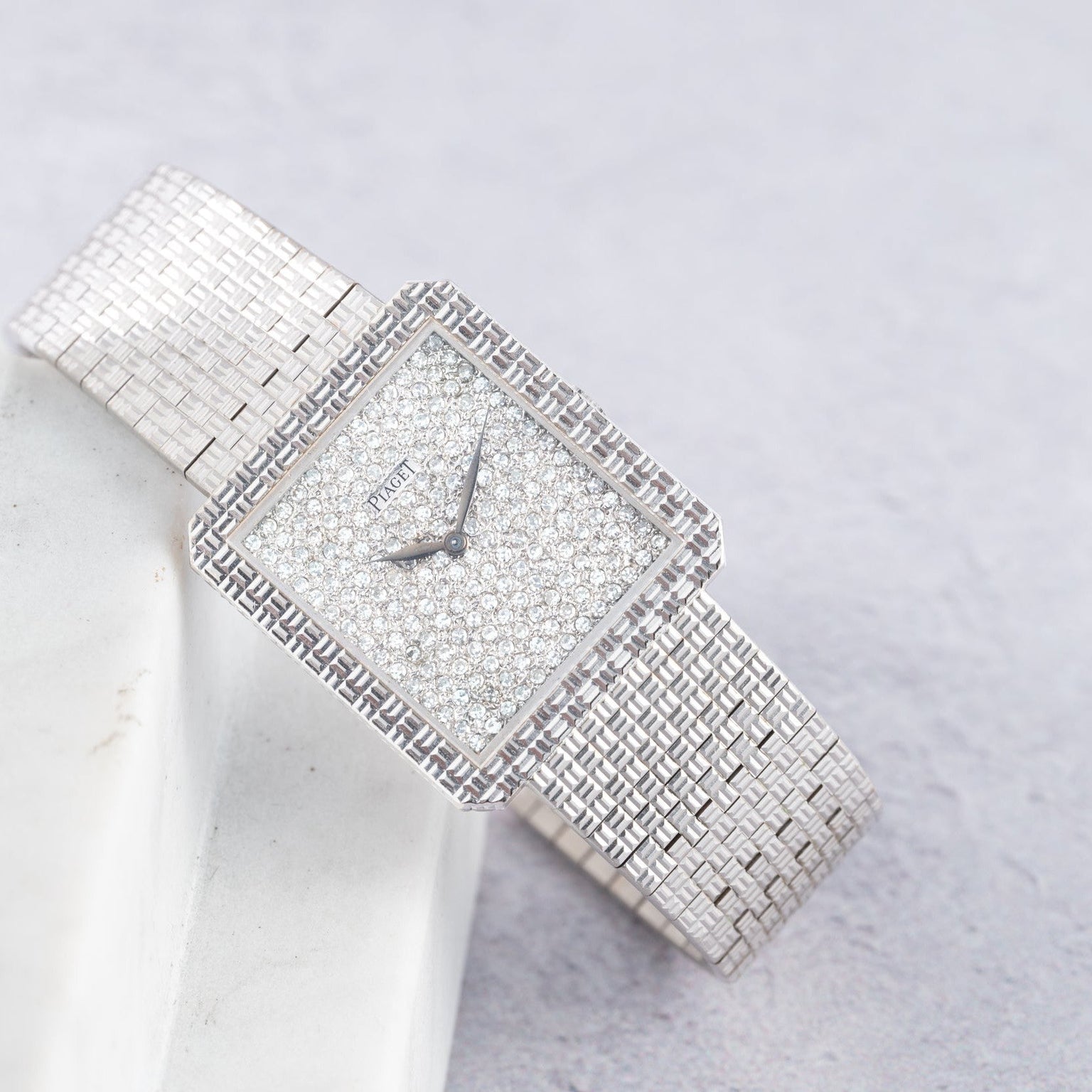 The Piaget Brick Diamond Dress Watch features a silver square face with a diamond-encrusted dial and band, elegantly displayed on a light marble surface.