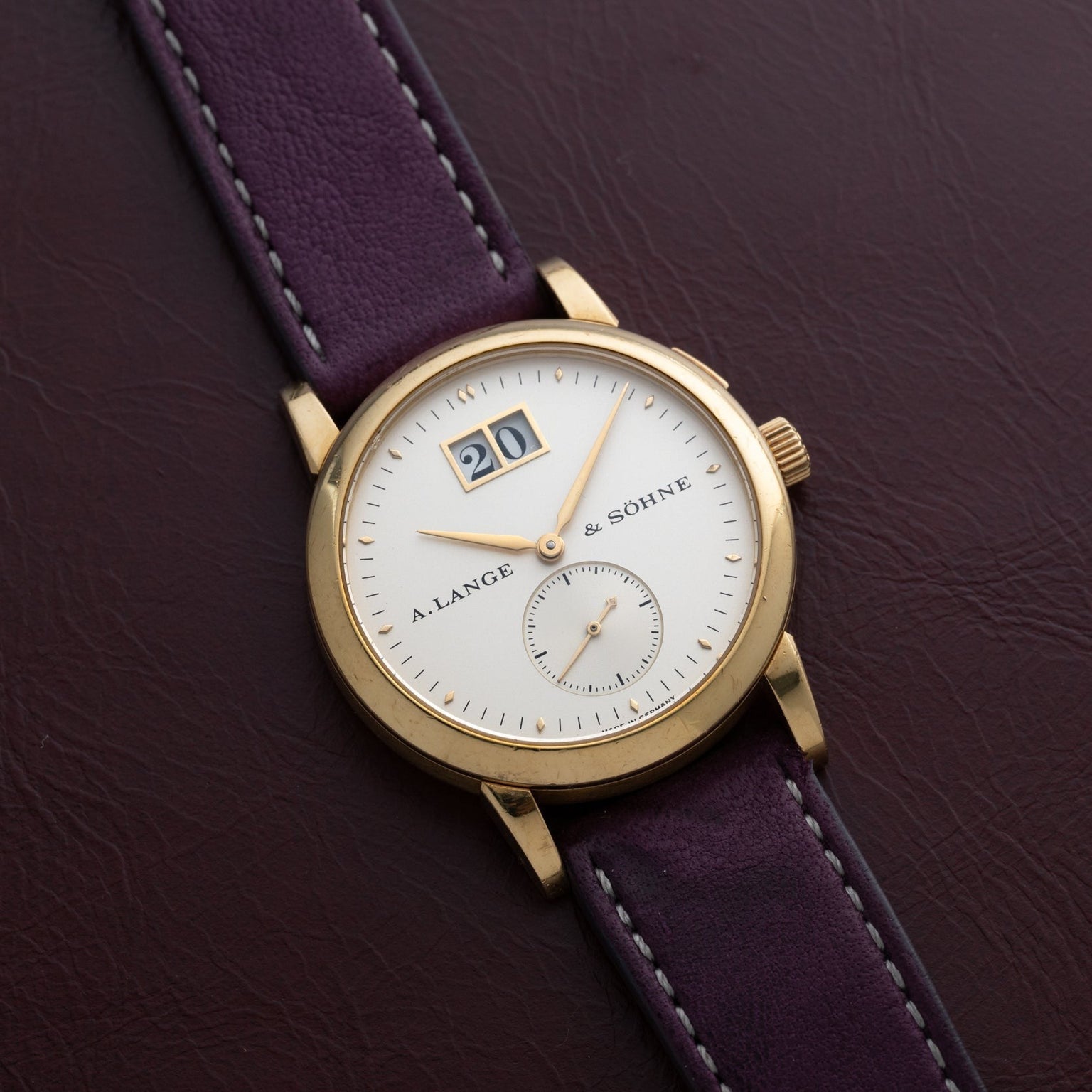 Lange & Sohne Saxonia Outsize Date wristwatch with a date display and purple leather strap placed on a brown leather surface.