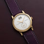 Lange & Sohne Saxonia Outsize Date wristwatch with a date display and purple leather strap placed on a brown leather surface.