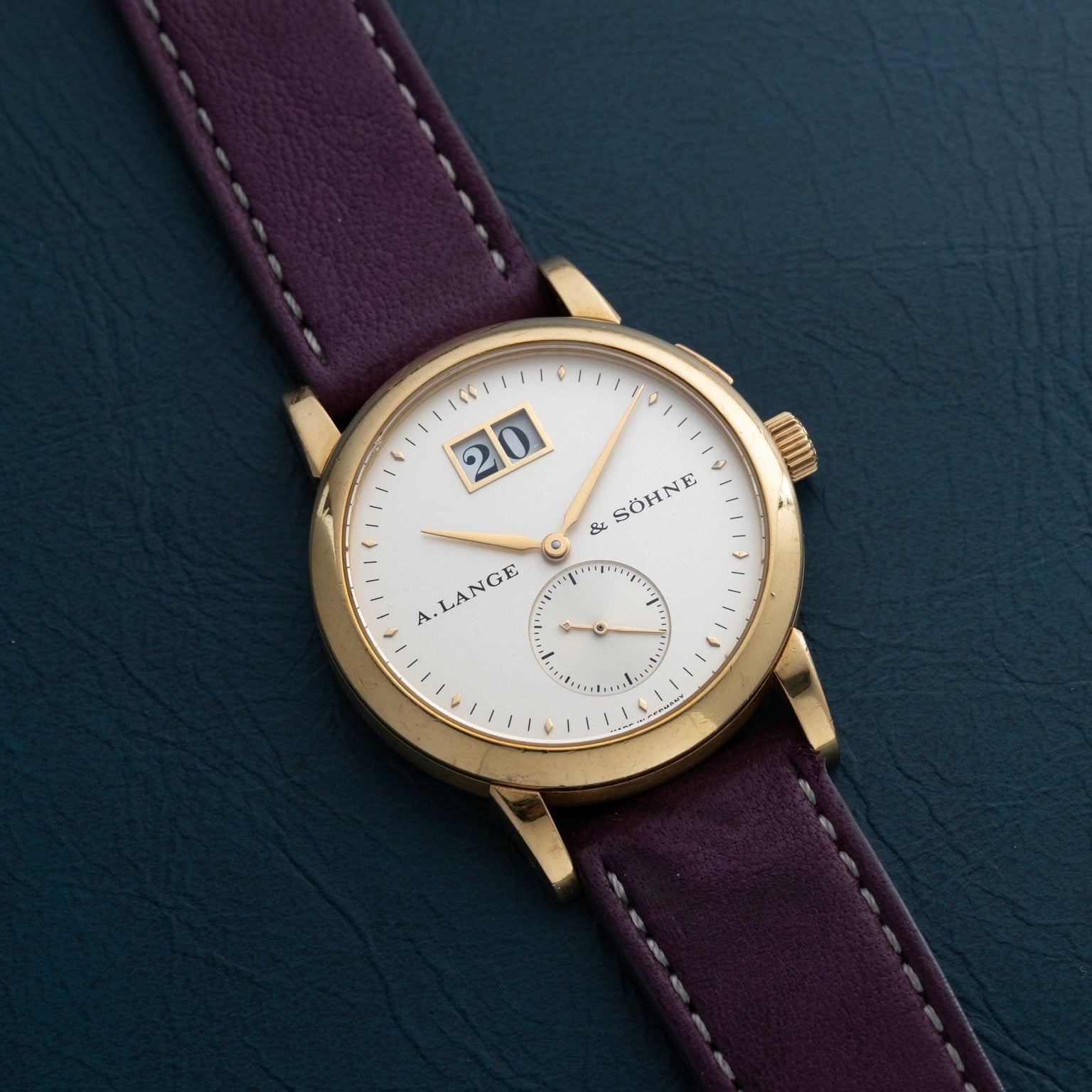 The Lange & Sohne Saxonia Outsize Date watch features a white dial, large date display, and purple strap, shown on a dark surface.